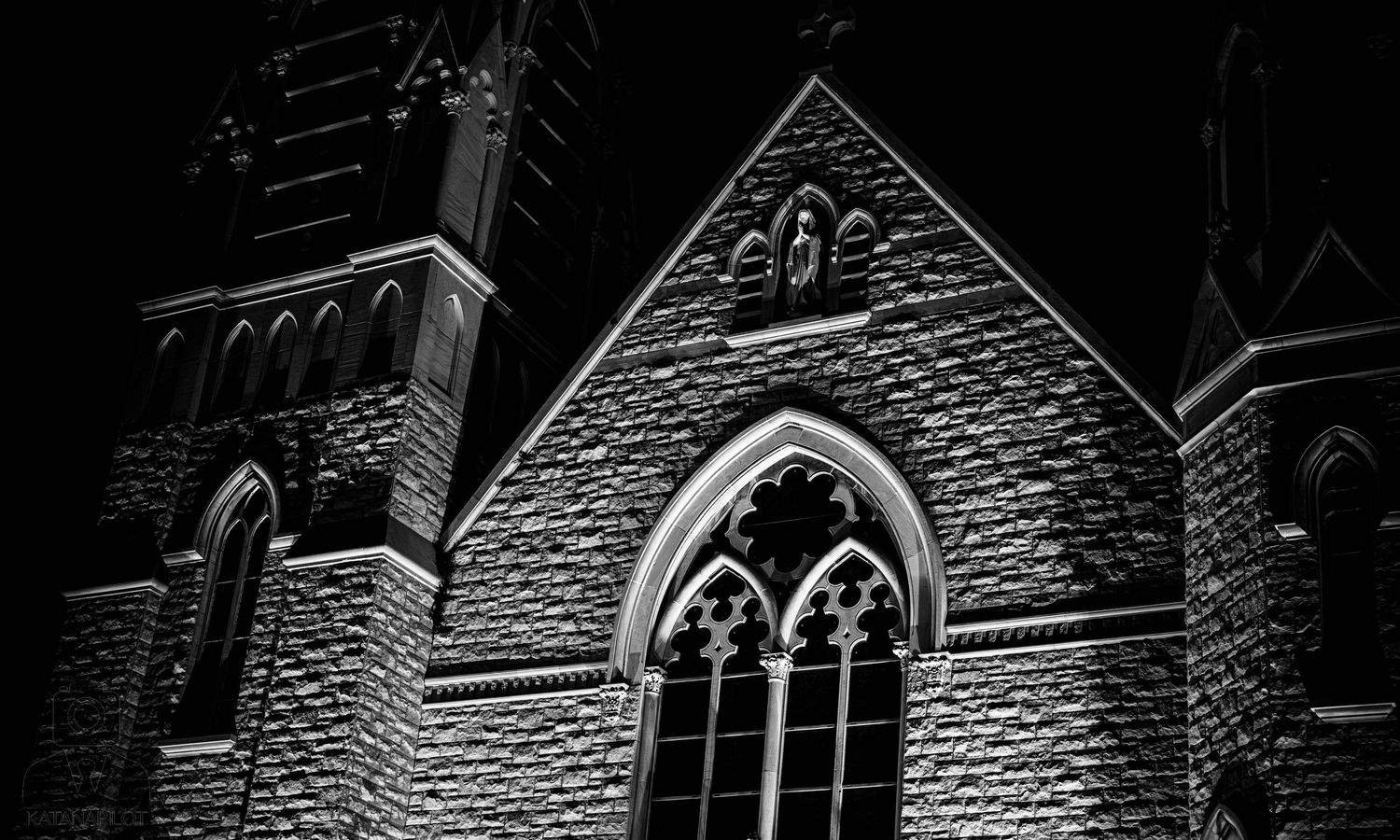 Black and white photo of a Gothic stone church at night with illuminated arched windows and ornate architectural details.