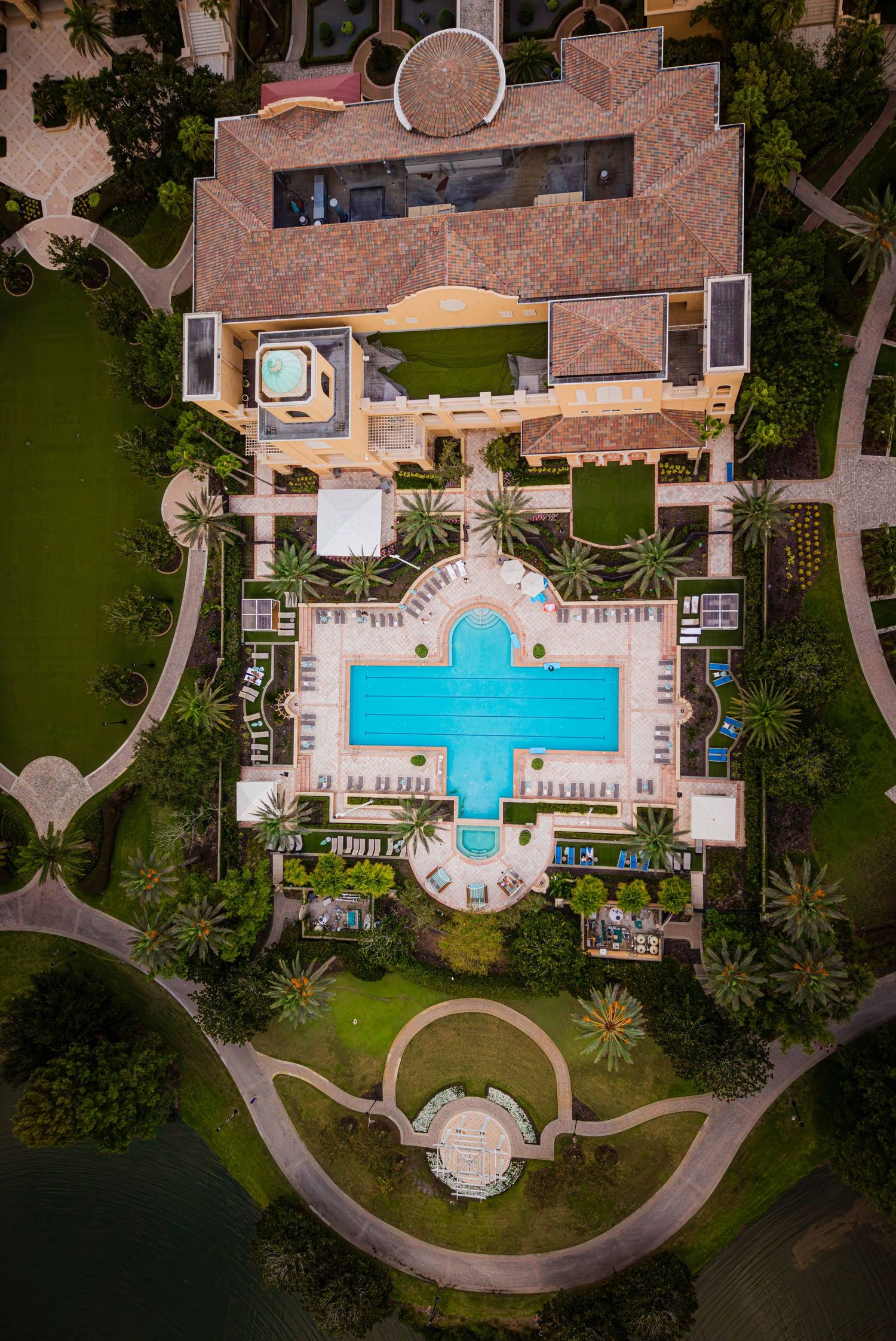 Aerial drone view of a luxury Mediterranean villa with a cross-shaped pool, manicured gardens, and palm trees.