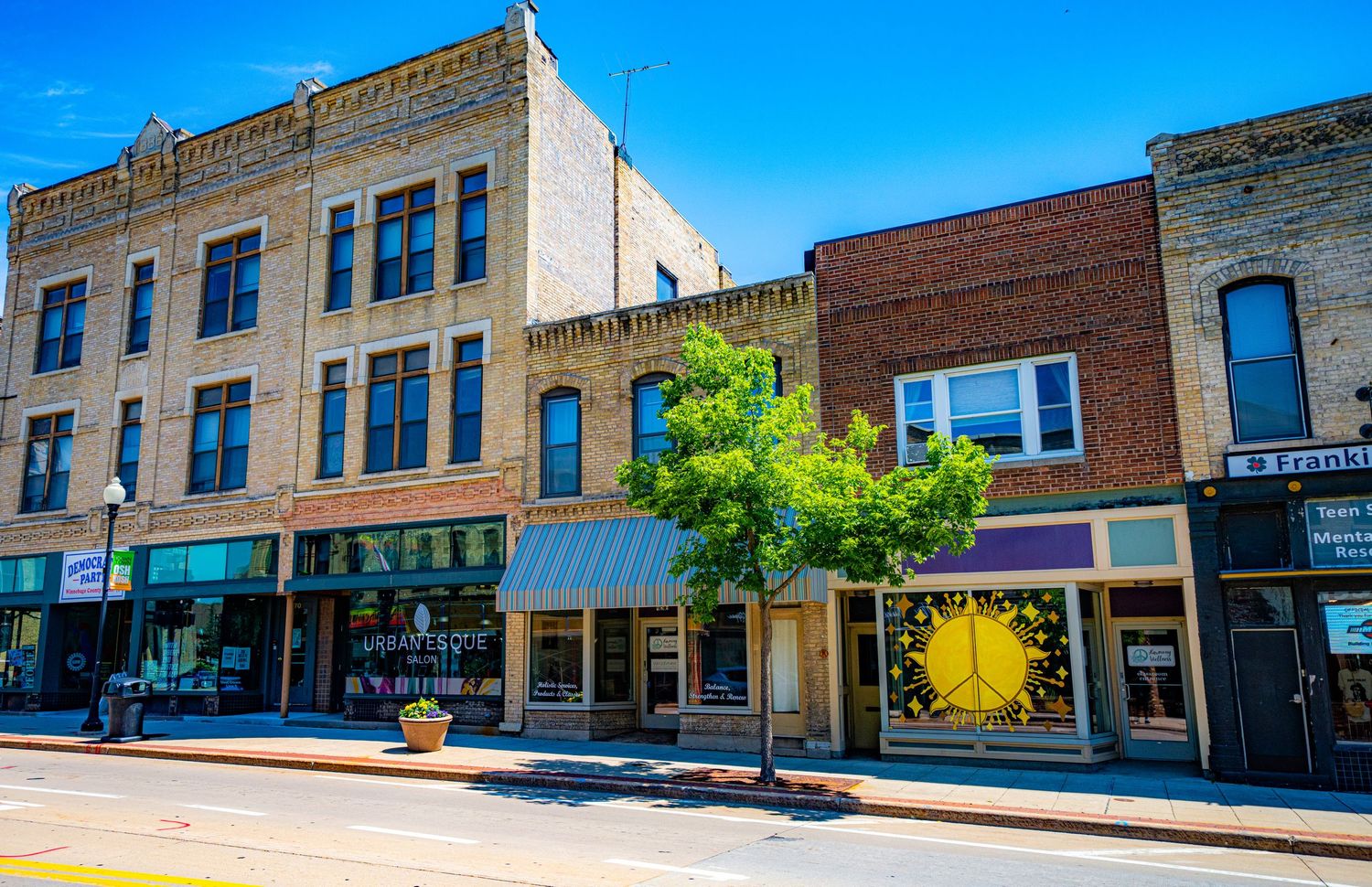Historic brick buildings with storefronts line a sunny main street in downtown Oshkosh.