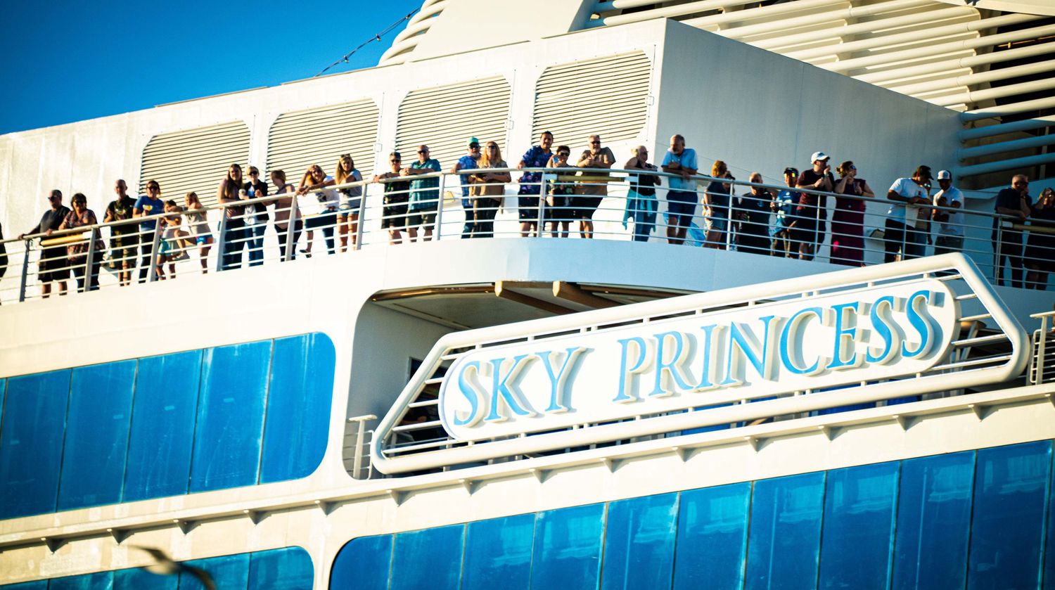 Passengers line the balcony deck of the Sky Princess cruise ship against a bright blue sky.