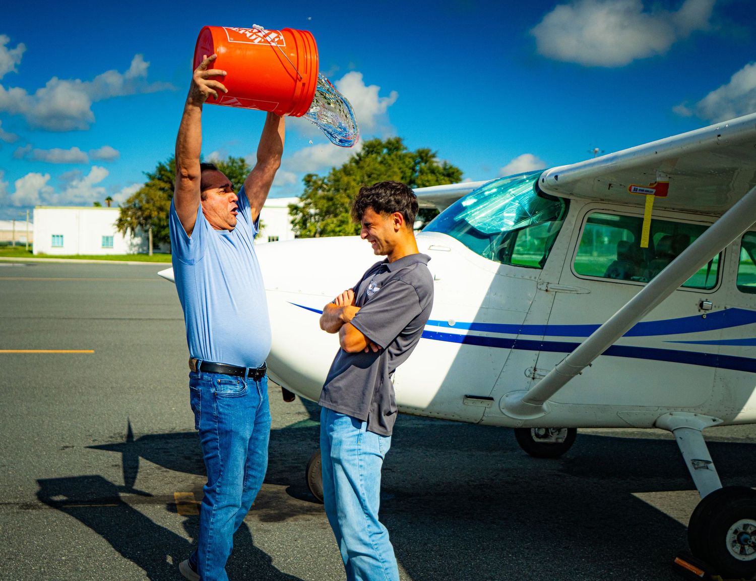 Man pouring fuel into a small Cessna aircraft on a sunny airport tarmac while a young student pilot observes.