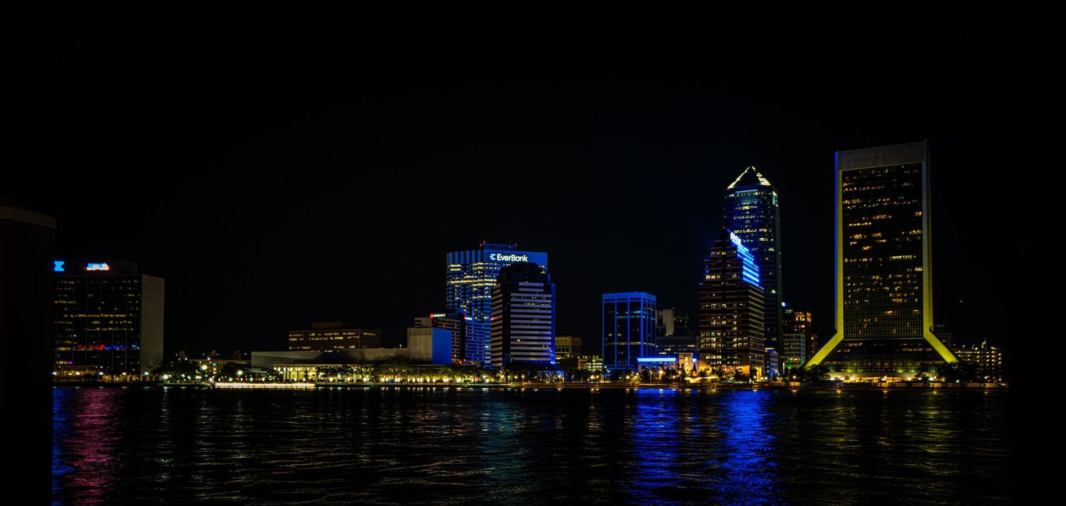 Jacksonville city skyline glowing with colorful lights reflected on the St. Johns River at night.