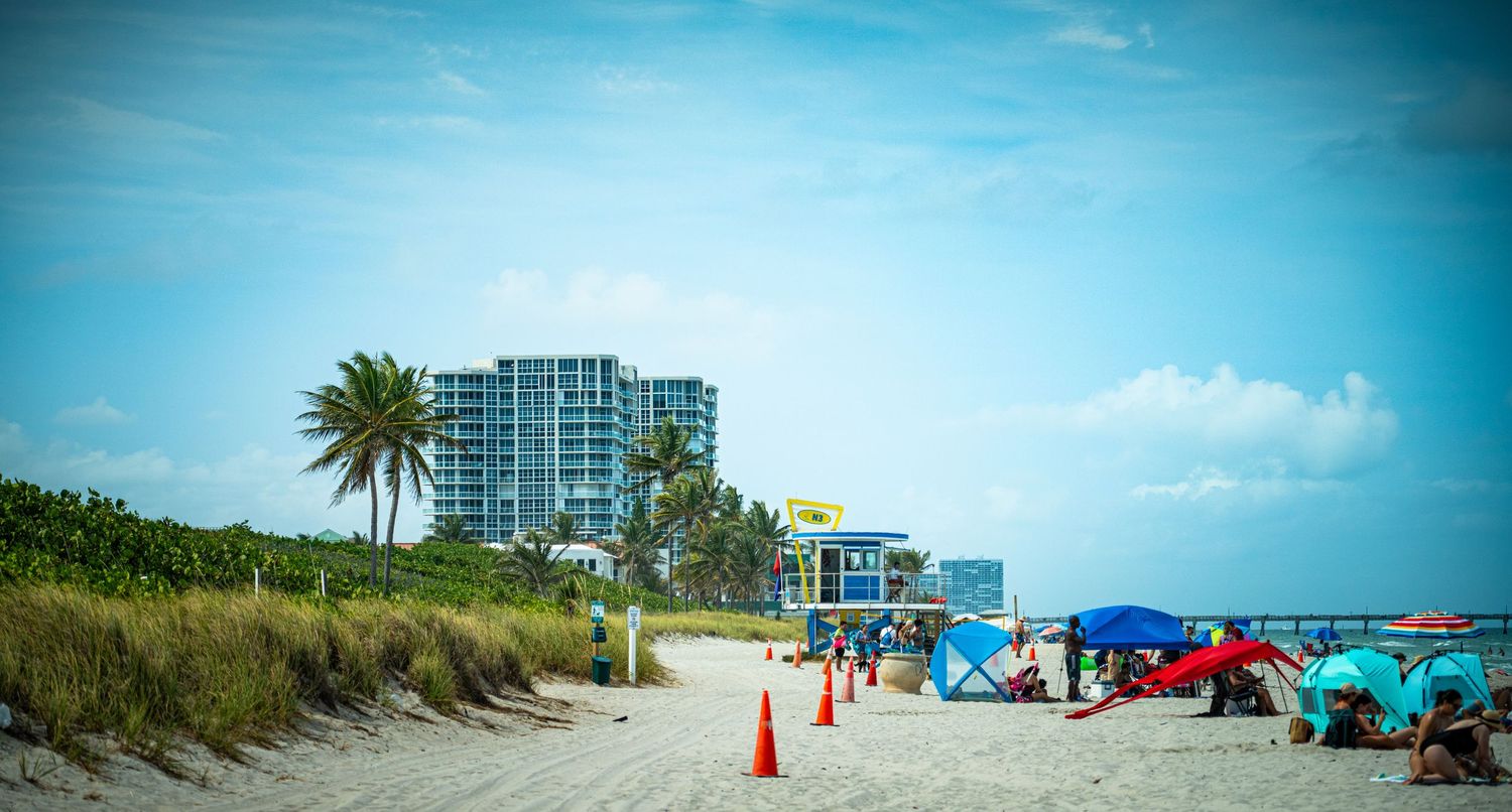 Panoramic view of a beach with high-rise buildings, palm trees, and scattered beach umbrellas.