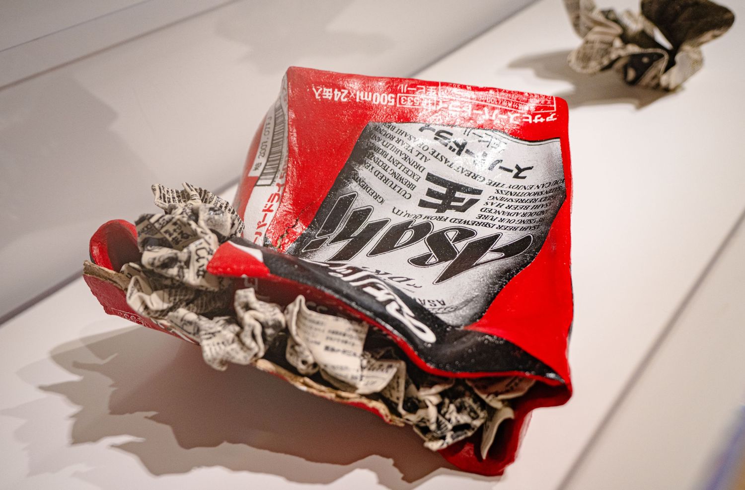 Crushed red Budweiser beer can art exhibit displayed on white surface in gallery.