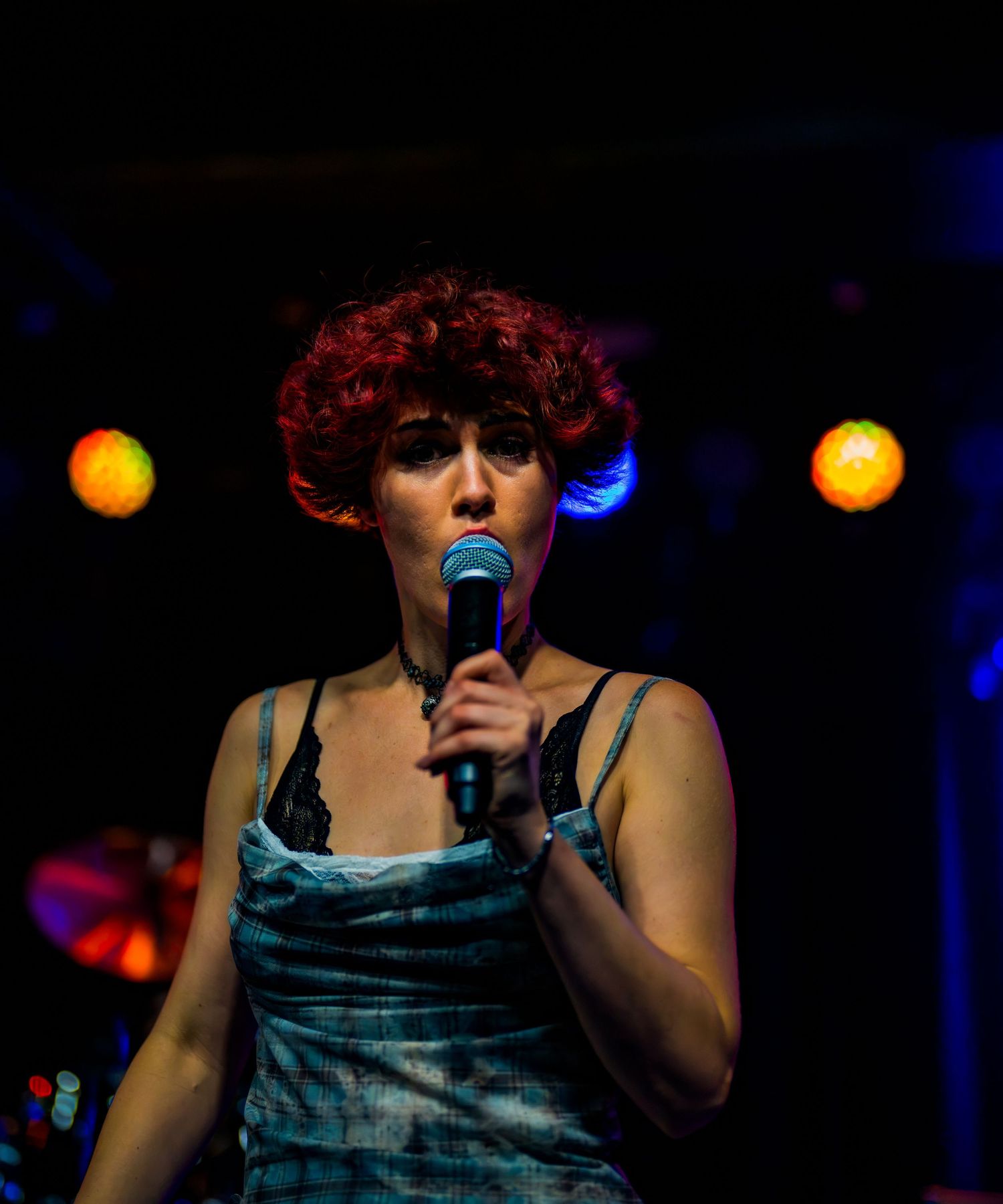 Female singer with red curly hair holding a microphone on a dark stage with colorful lights.