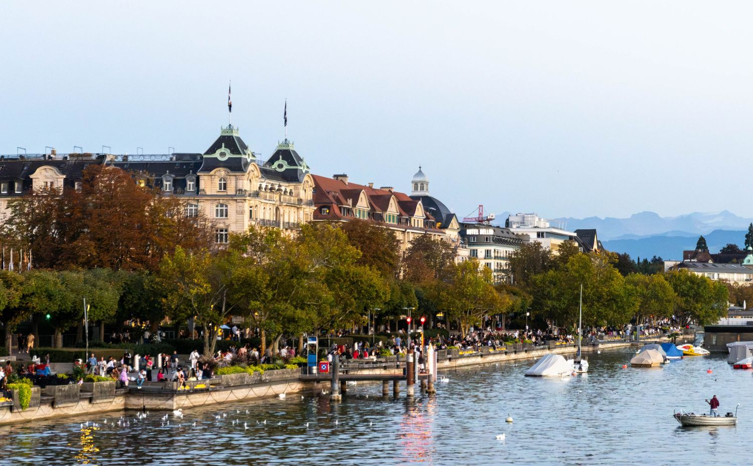 Lakeside promenade with autumn trees, historic buildings and boats on calm water, with hazy mountains rising in the distance.