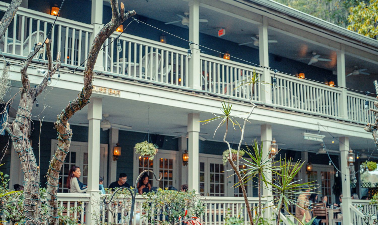 A historic white wooden building with a wraparound porch and balcony features tropical landscaping and palm trees.