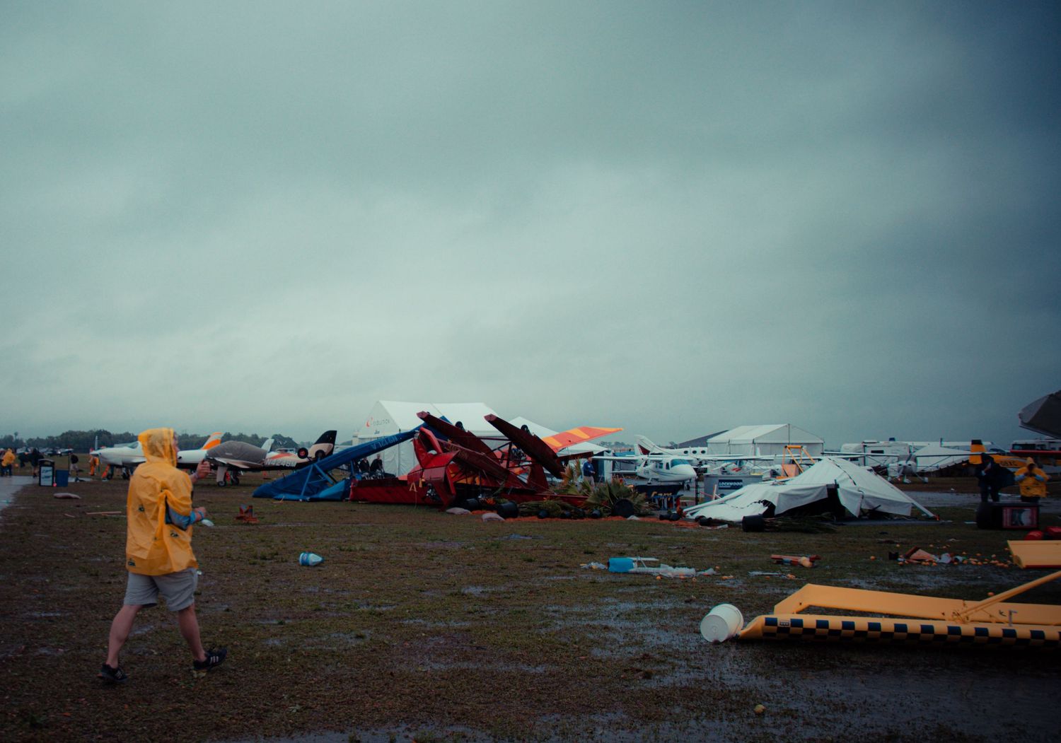 A figure in a yellow raincoat walks past storm damage and debris from destroyed aircraft at an airfield.