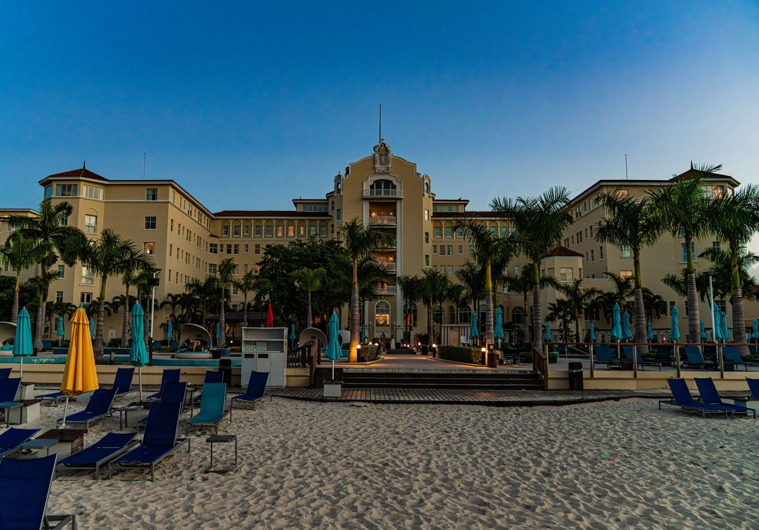 Beachfront luxury hotel with palm trees and blue lounge chairs on white sand at dusk.
