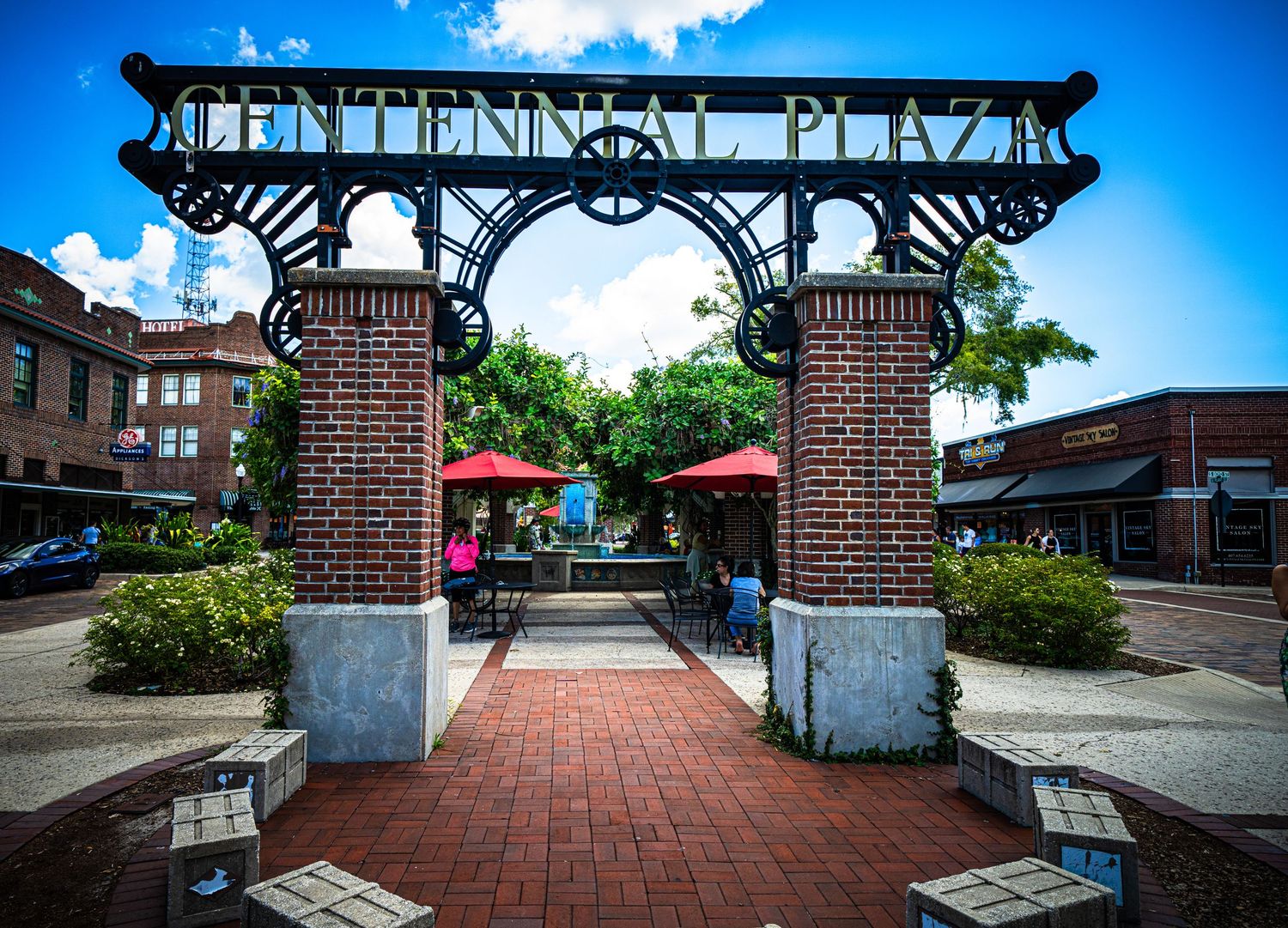Decorative iron archway entrance with brick pillars leading to commercial plaza.