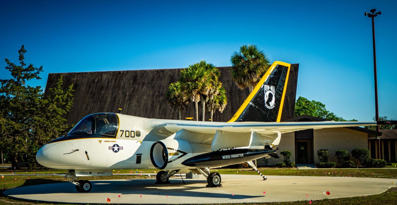 A vintage US Navy S-3 Viking jet aircraft on outdoor display near a building with palm trees under a clear blue sky.