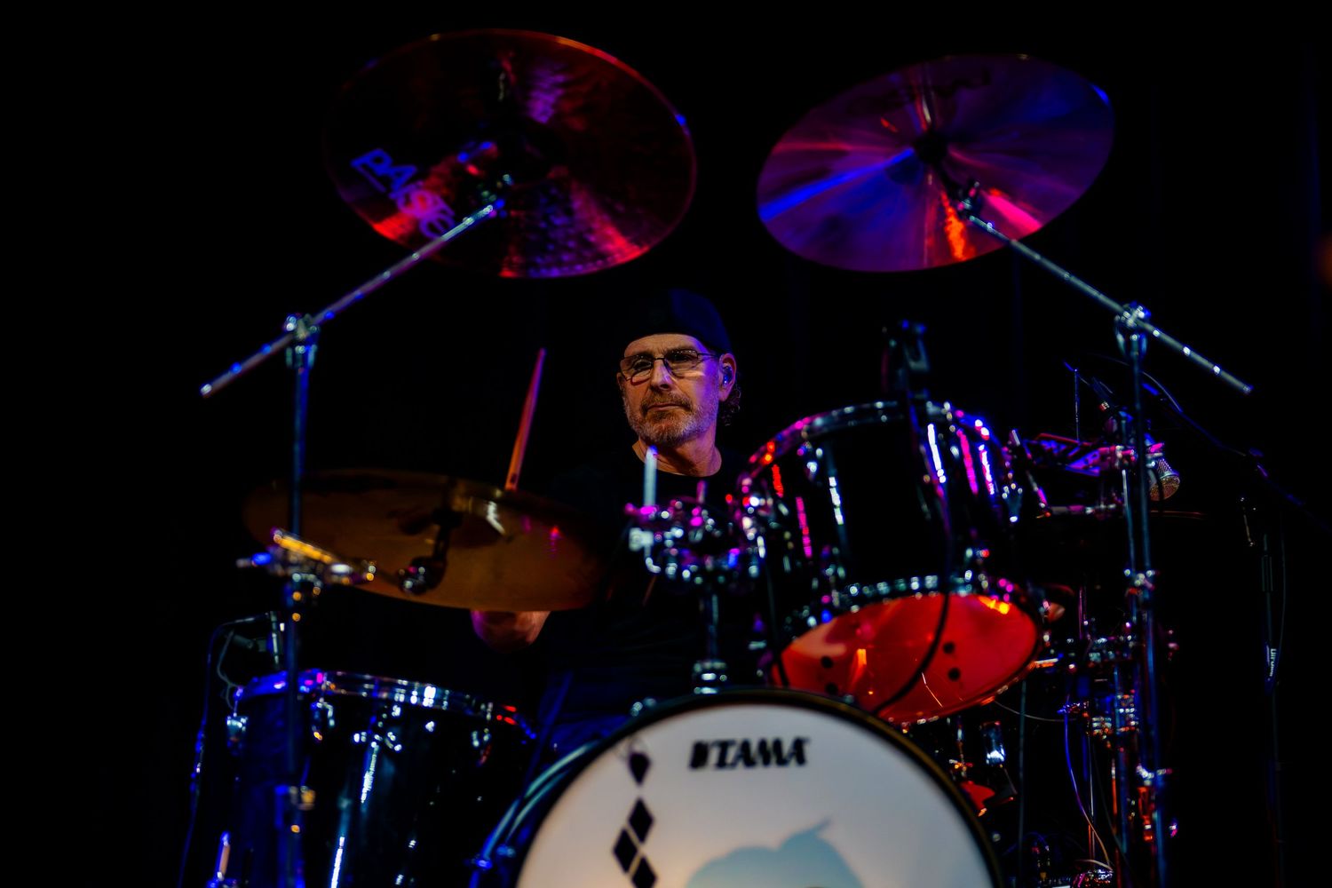 Musician playing Tama drum kit on stage with illuminated cymbals in dramatic concert lighting.