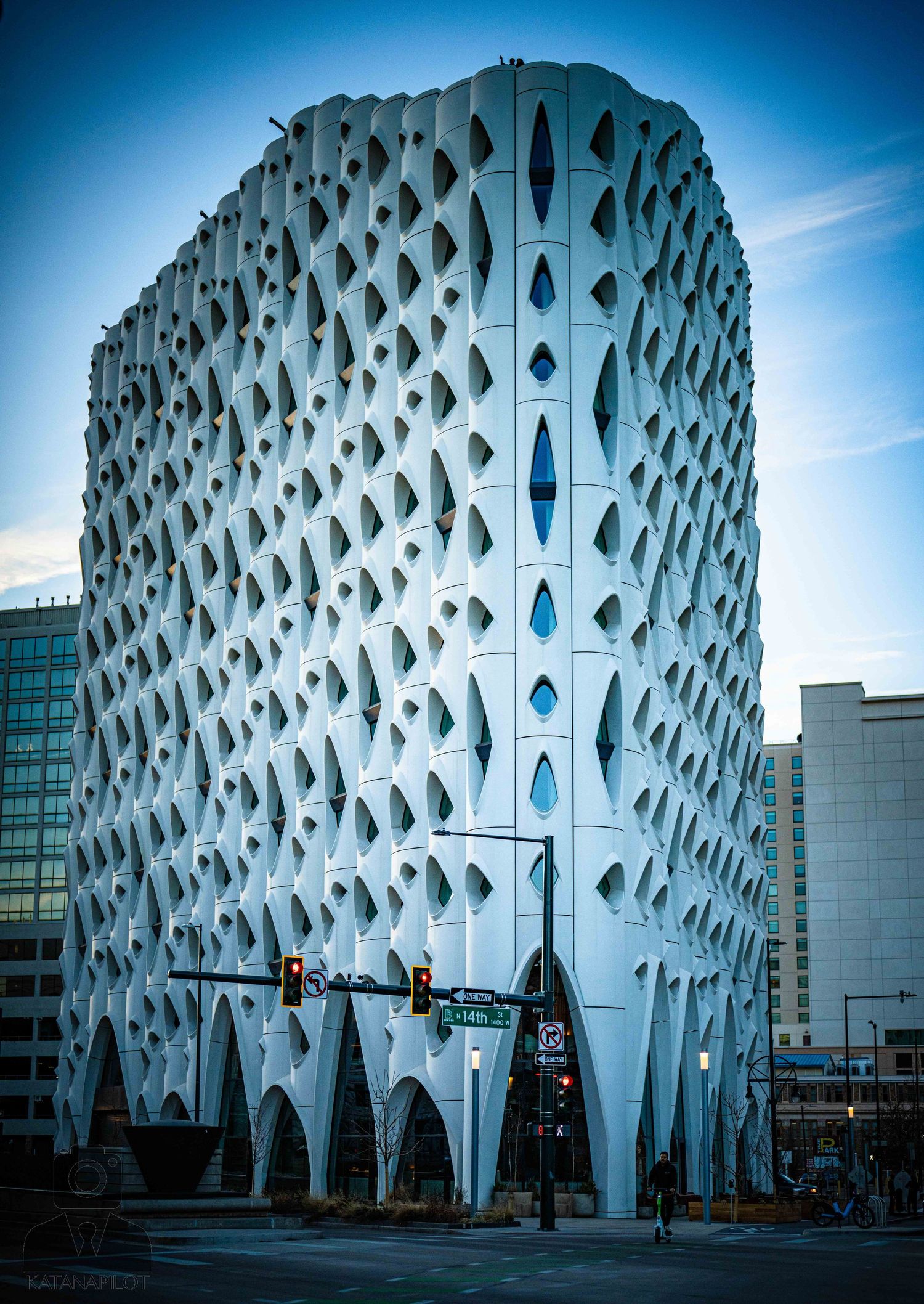 Modern architectural building with honeycomb-like perforated white facade and arched base in an urban setting against blue sky.