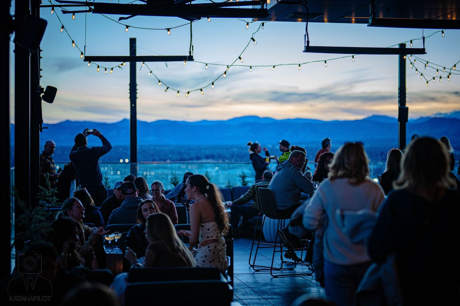People gather at a scenic mountain viewpoint with chairs and tables overlooking a dramatic sunset landscape.