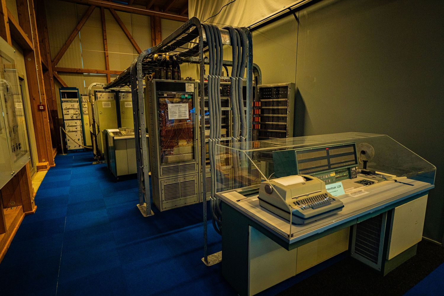 Vintage mainframe computer cabinets with bundled cables and a control console with printer on display in a museum room with blue carpet.