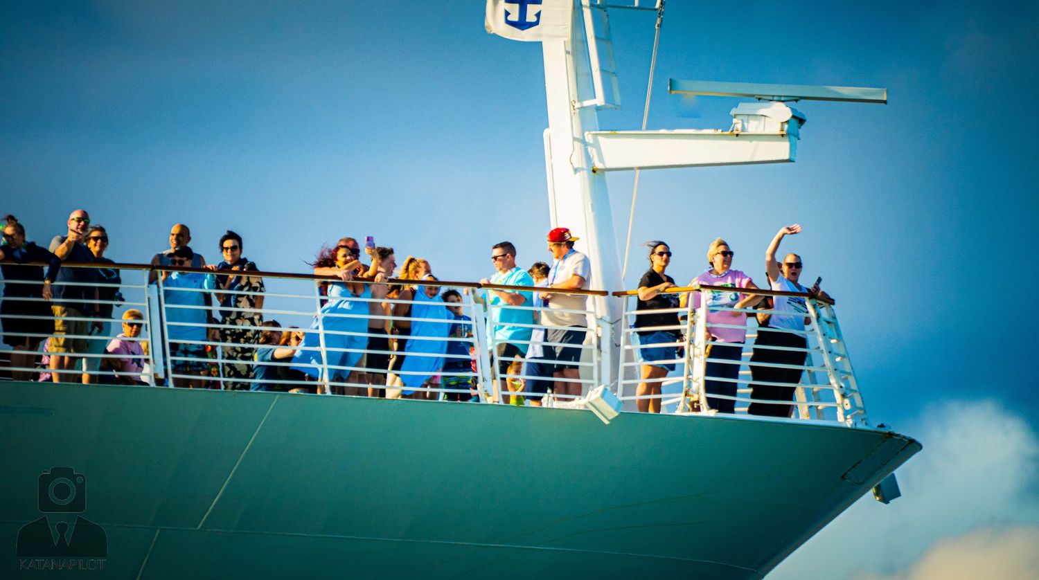 Passengers line the upper deck railing of a cruise ship sailing on bright blue waters.