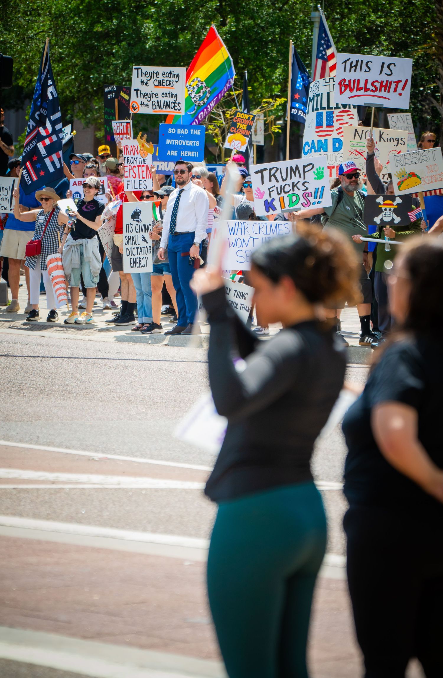 Protesters hold colorful signs and rainbow flags at a political demonstration along a street.