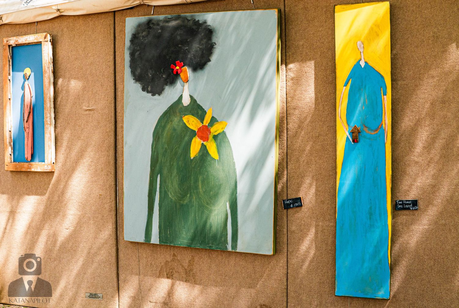 Series of colorful paintings displayed on tan walls at an outdoor art exhibition.