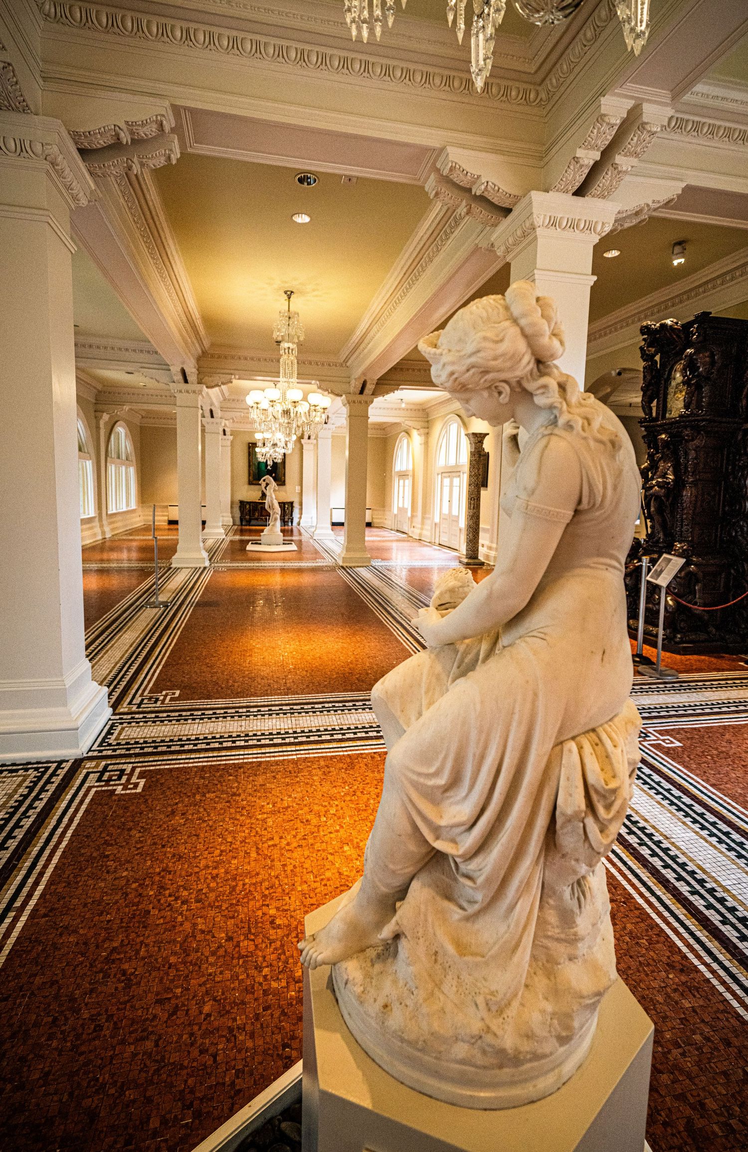 Grand hallway with classical marble statues, crystal chandeliers and decorative orange carpeting.