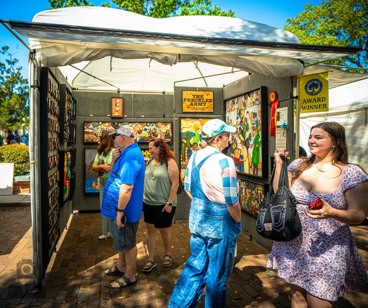 Visitors browse merchandise at an outdoor market stall with white canopy on a sunny day.