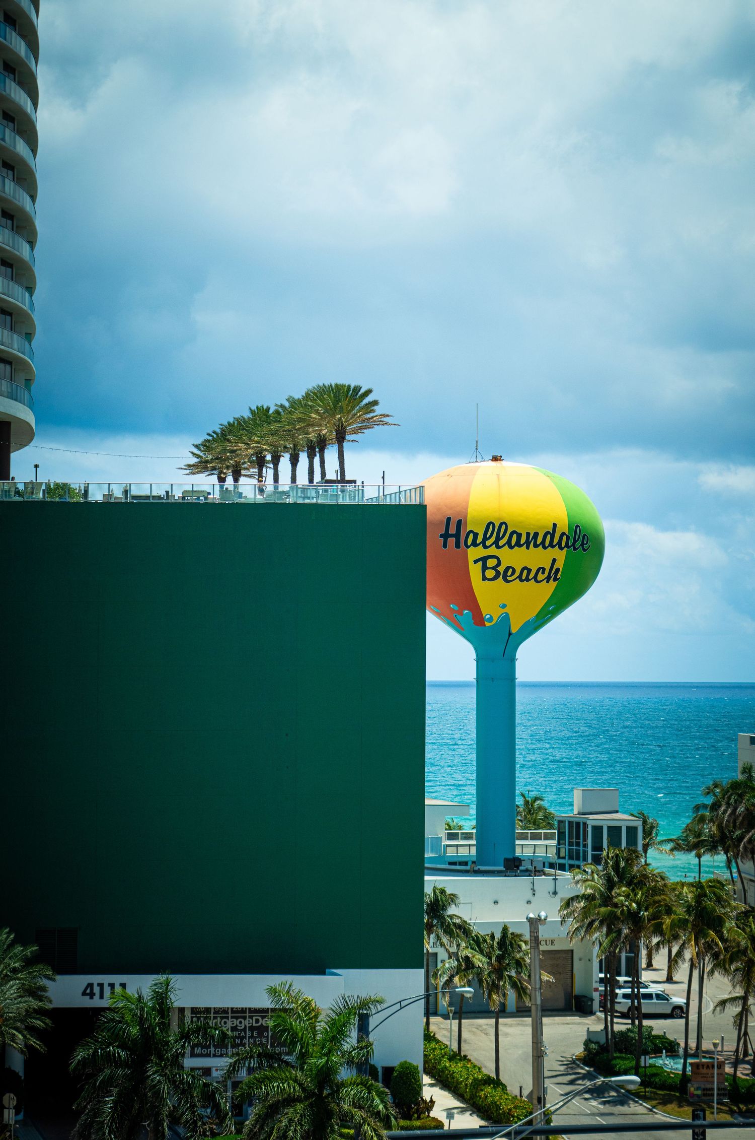 Oceanfront view of palm trees and high-rise buildings with a distinctive water tower against a cloudy blue sky.
