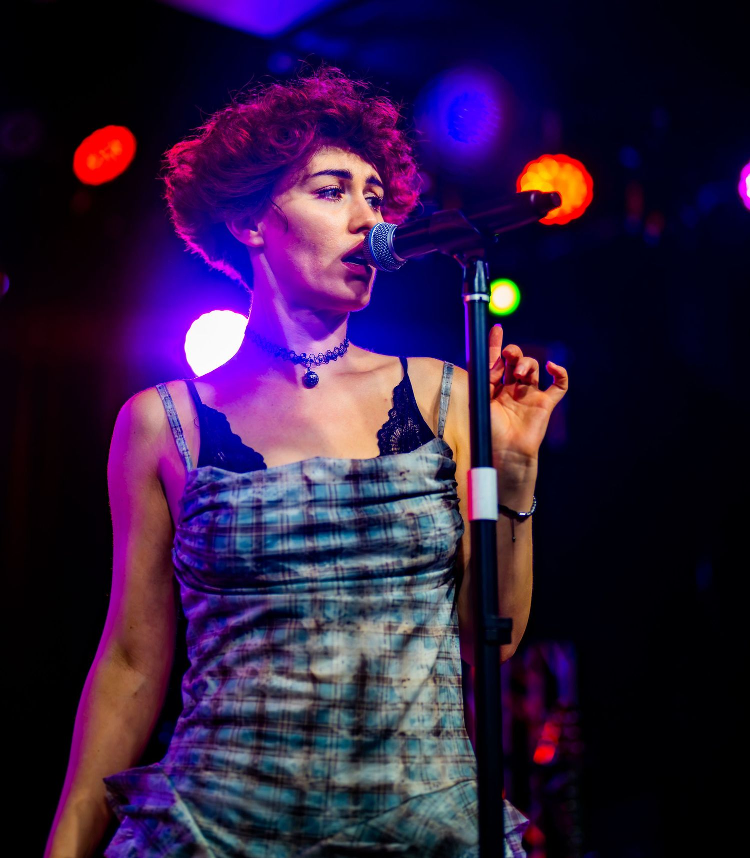 Female vocalist with curly red hair sings into microphone wearing choker necklace on a lit concert stage.