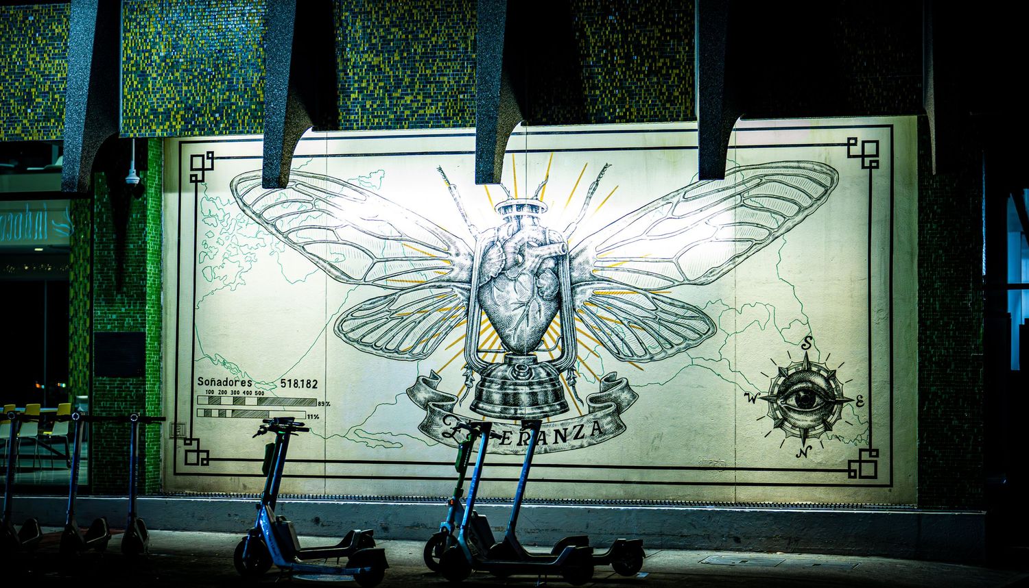 Vibrant street mural featuring a heart with butterfly wings and compass on a building wall, with electric scooters parked in front.