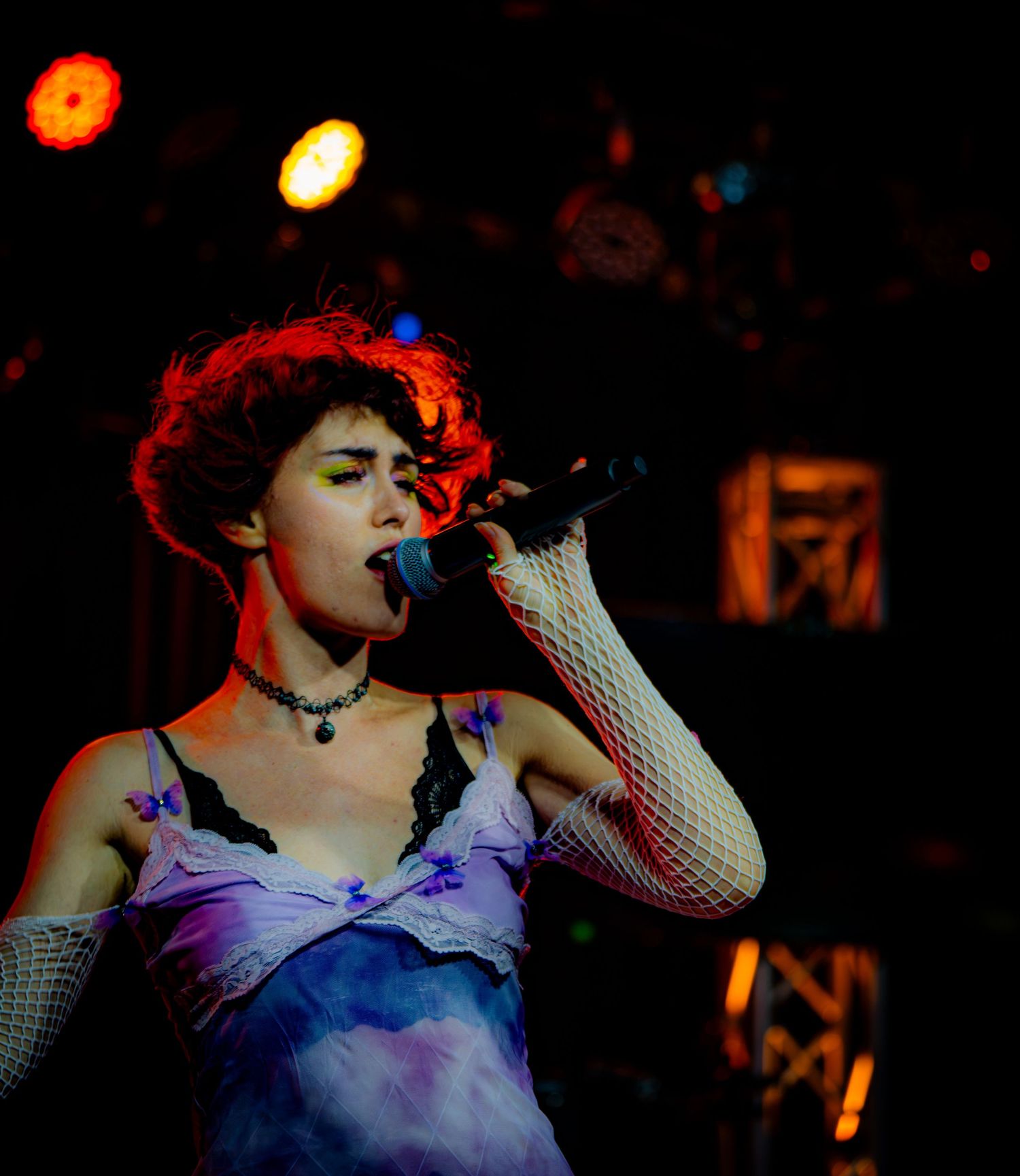 Young female singer performing on stage, wearing fishnet gloves and floral dress, singing into microphone under red stage lights.