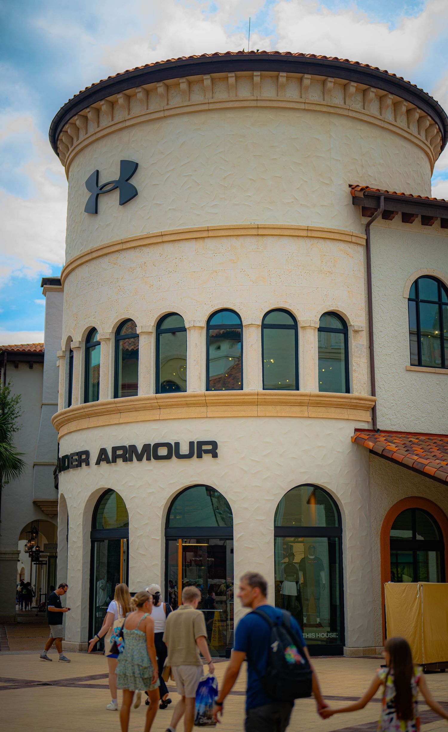 Under Armour retail store exterior with rounded architecture and logo, shoppers walking outside on a sunny day.