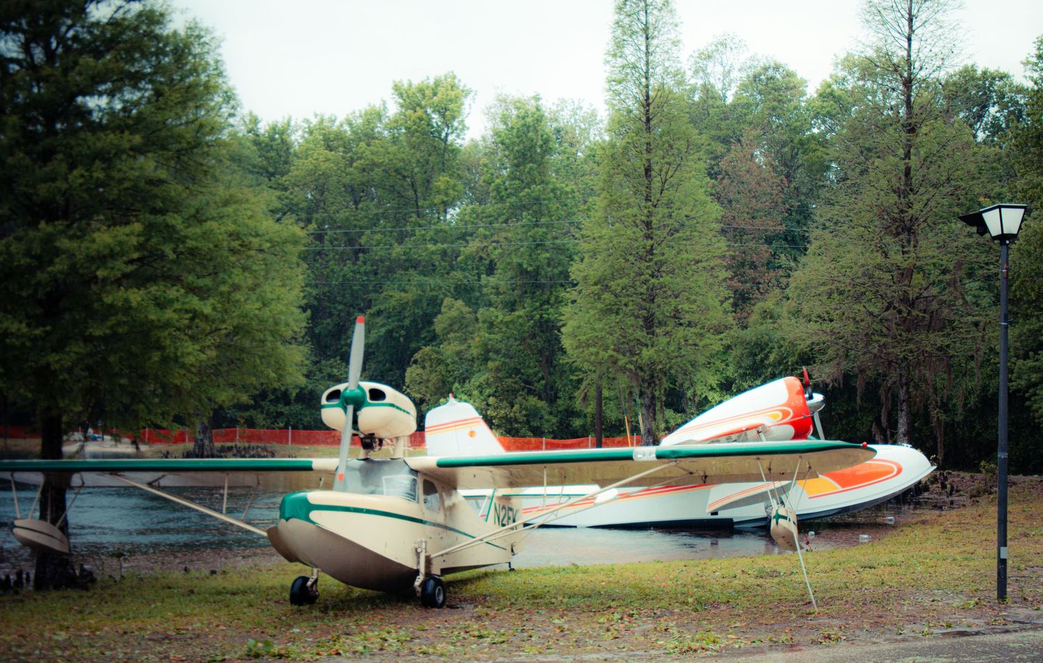 A vintage seaplane with red and white paint scheme sits parked among pine trees in a forest setting.