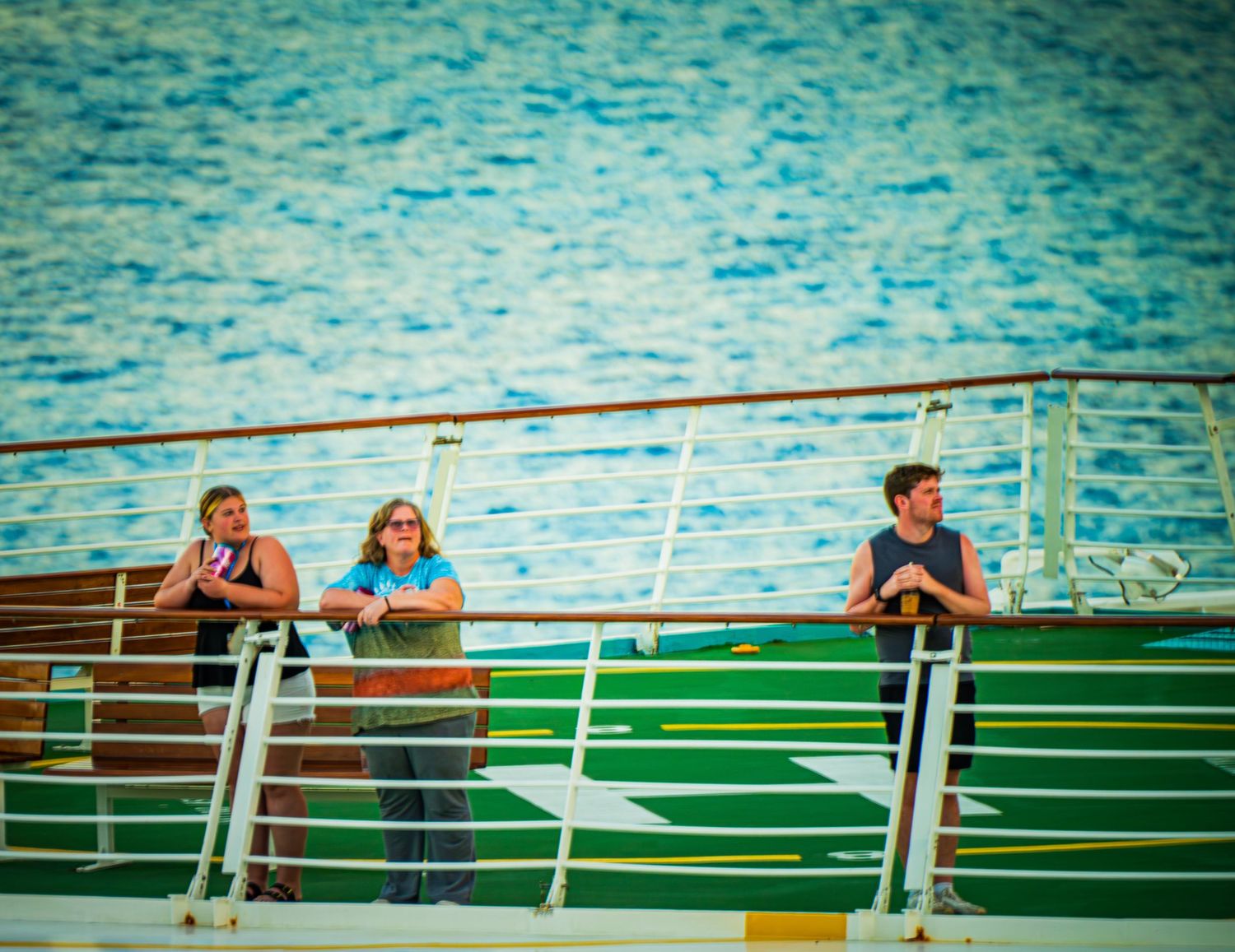People lean against a railing overlooking turquoise water on a sunny day at sea.