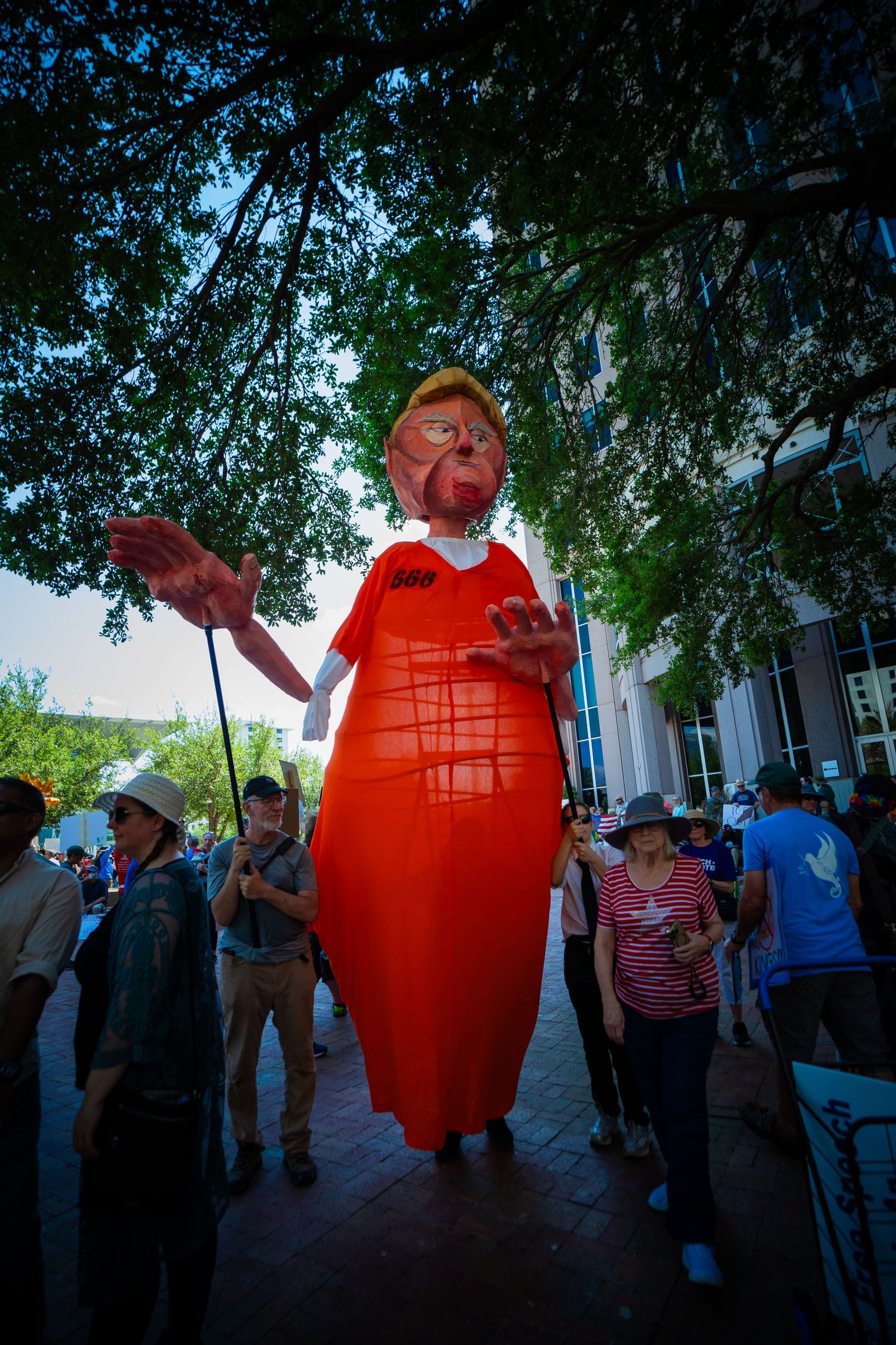 A large inflatable figure in a bright red dress stands among a crowd of people under tree branches.