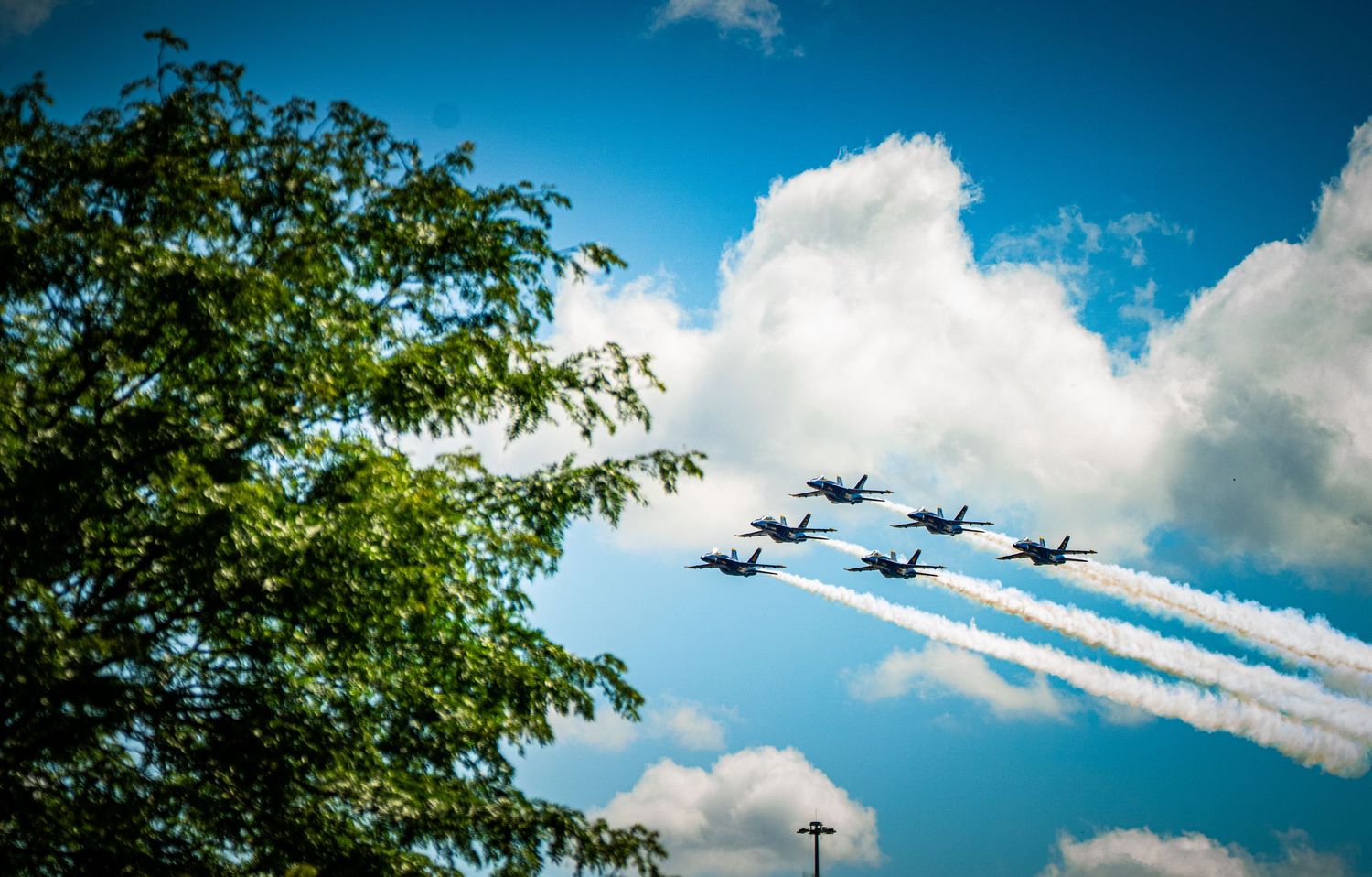 Military aircraft performing aerial display with contrails against blue sky and white clouds.