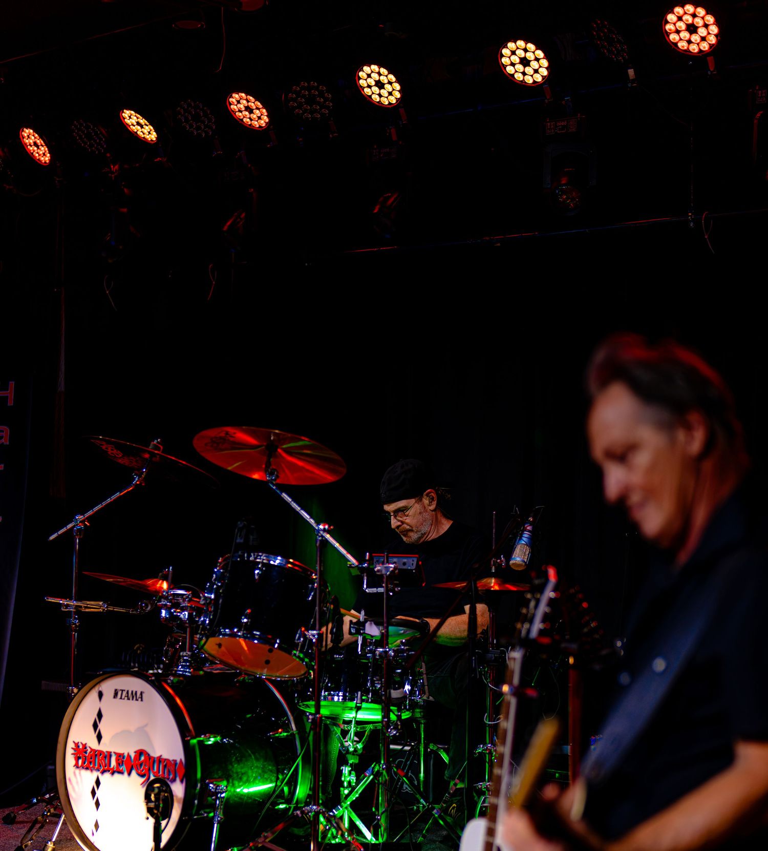 Live band performance with drummer on stage and guitarist in foreground under colorful concert lights.