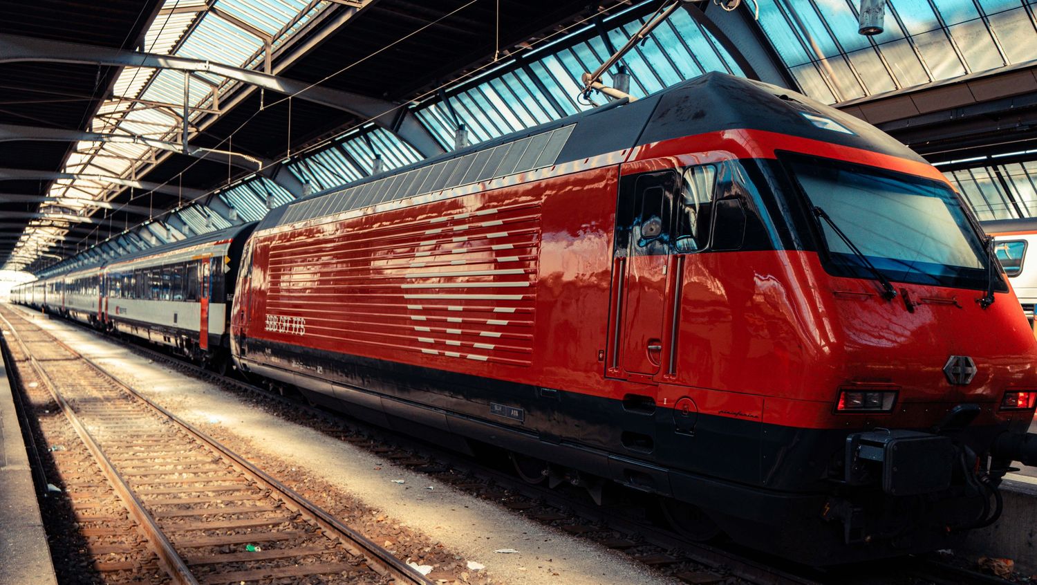 Red electric passenger train with white stripes stopped at a covered railway station platform under a glass and steel roof.