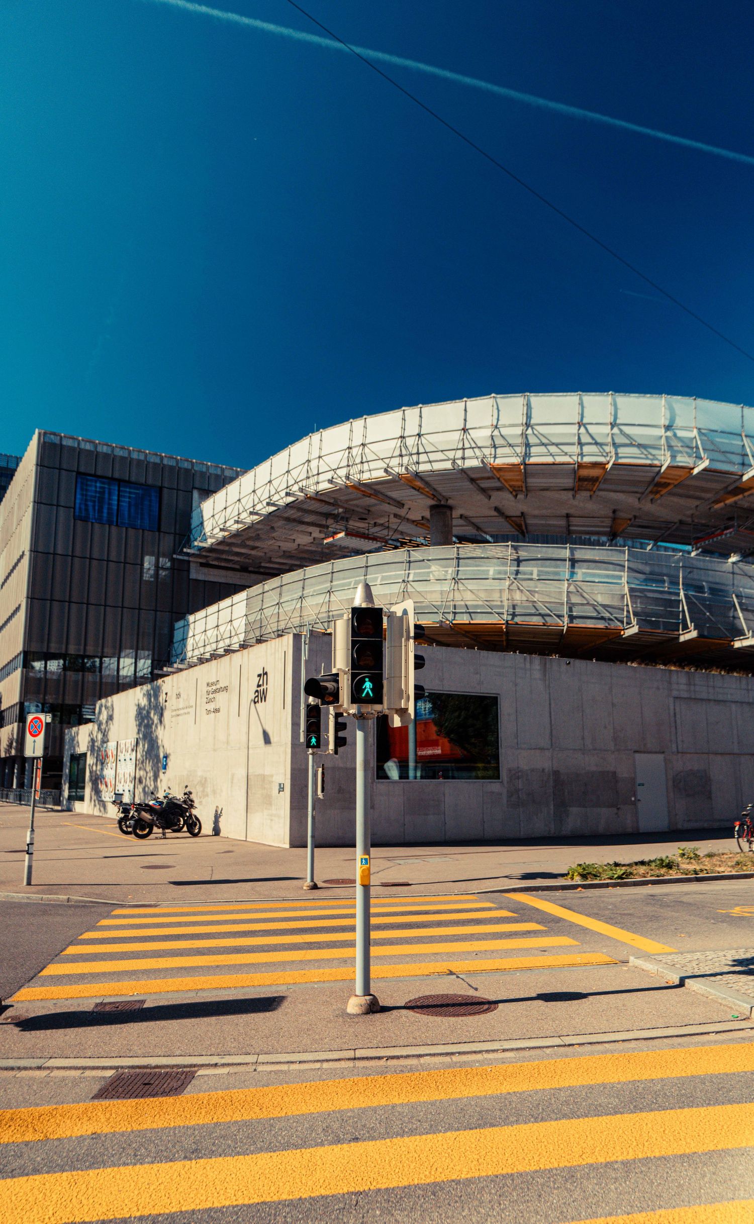 Modern curved glass and concrete building beside a pedestrian crossing with yellow stripes and a green traffic light under a deep blue sky.