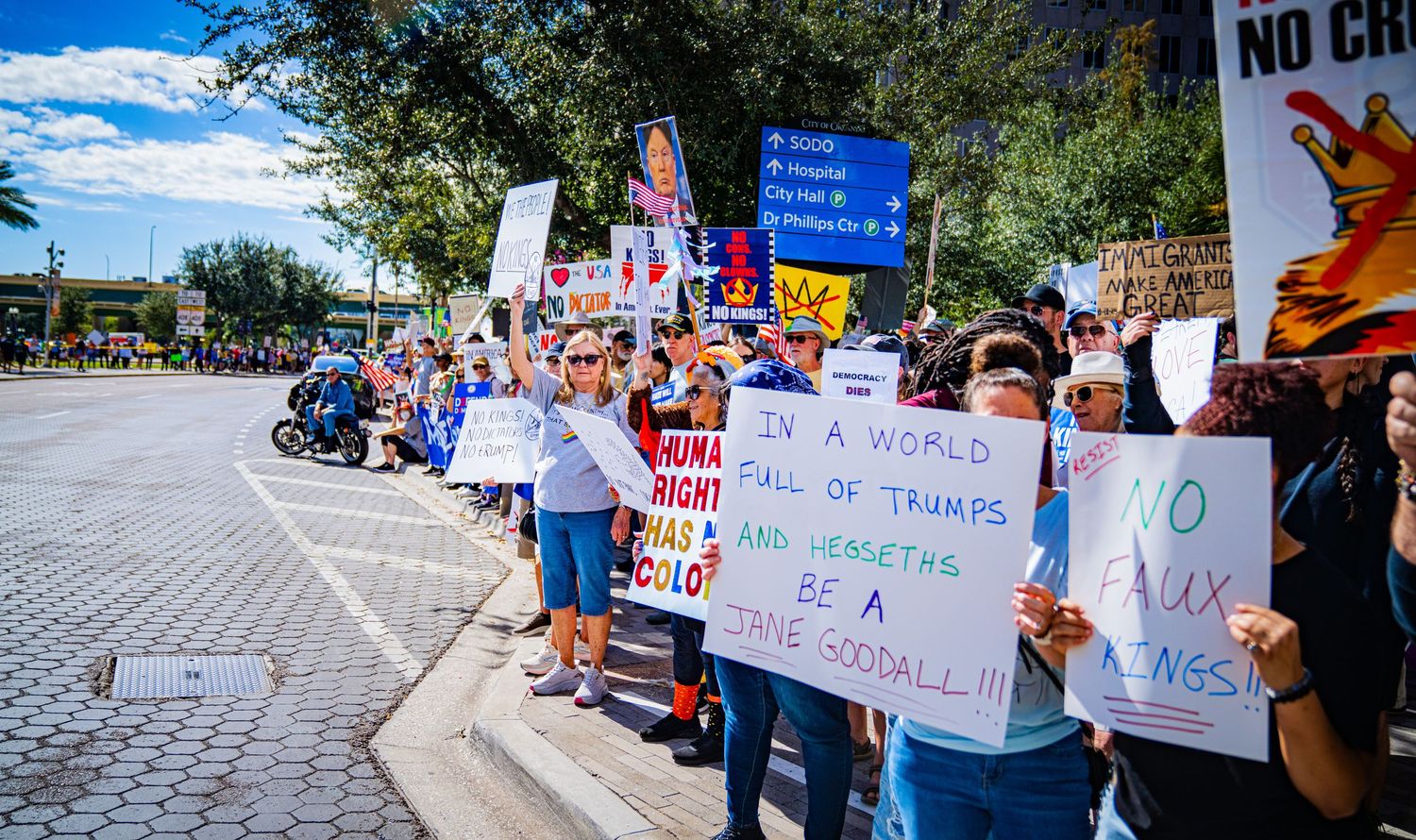 Protesters gather on a sidewalk holding signs and banners during a demonstration on a sunny day.