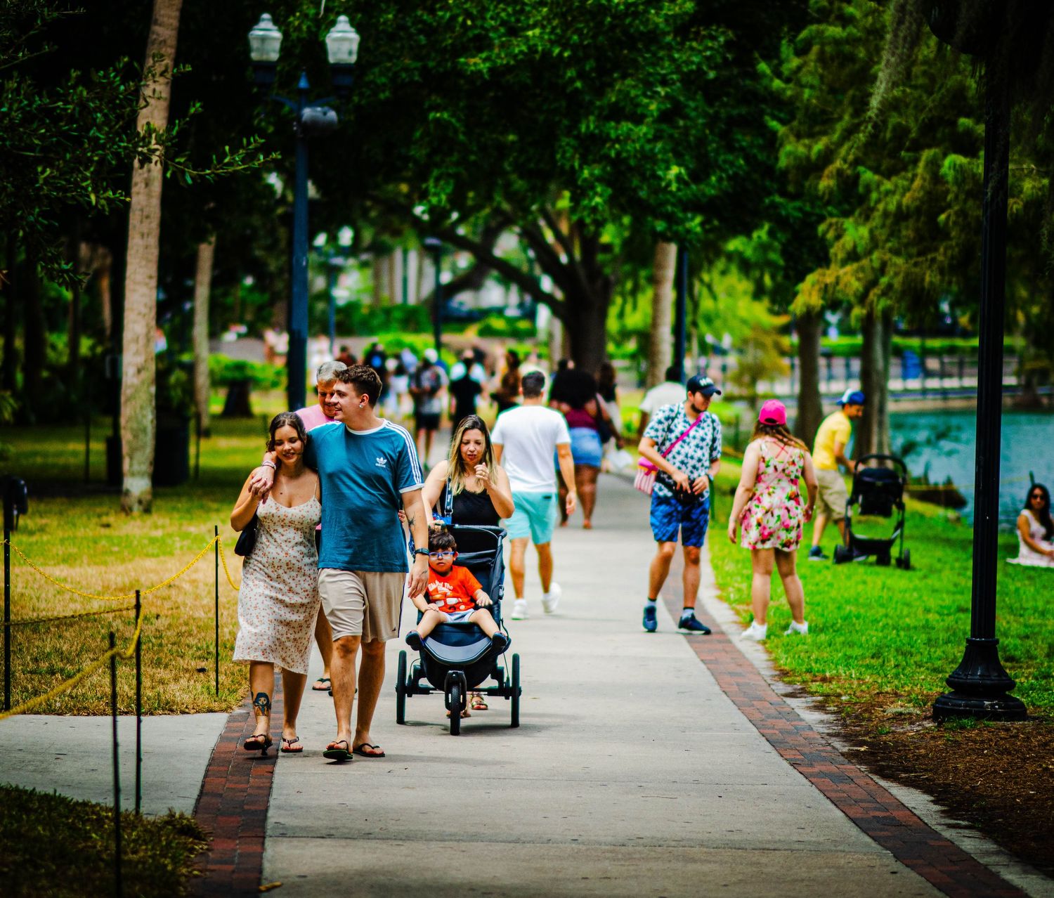 Families and couples stroll along a sunny park walkway lined with palm trees near a waterfront.