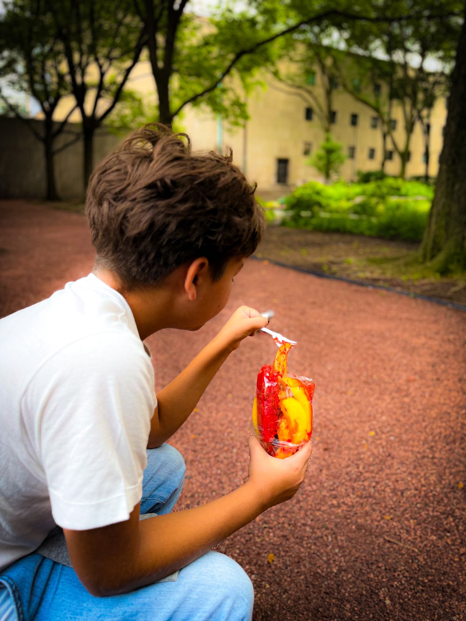 A boy crouching outdoors, drinking from a colorful red and yellow beverage pouch with a straw.
