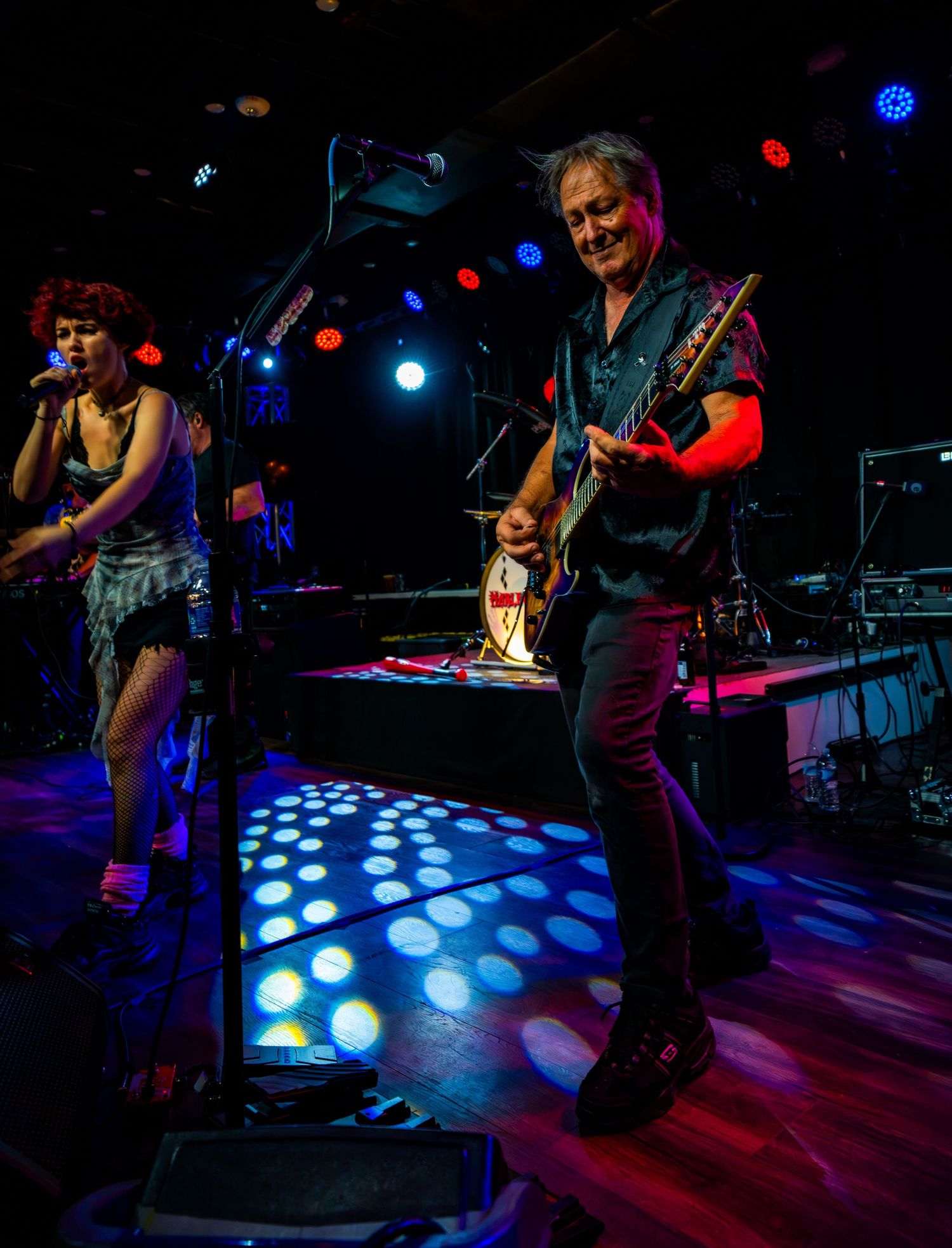 Female singer and male guitarist performing together on colorful disco-lit concert stage.