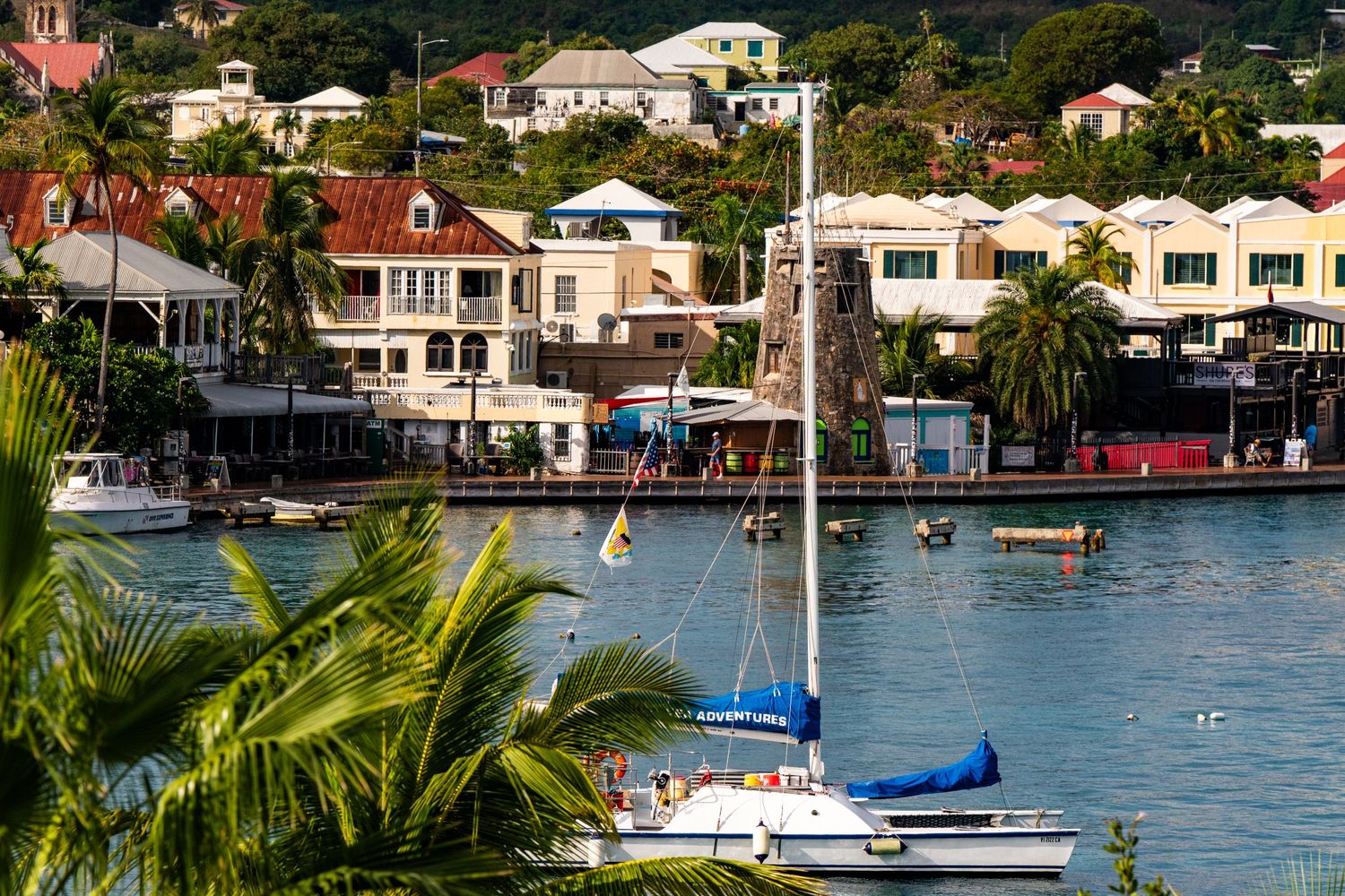 Tropical harbor scene with sailboats and waterfront buildings under blue skies.
