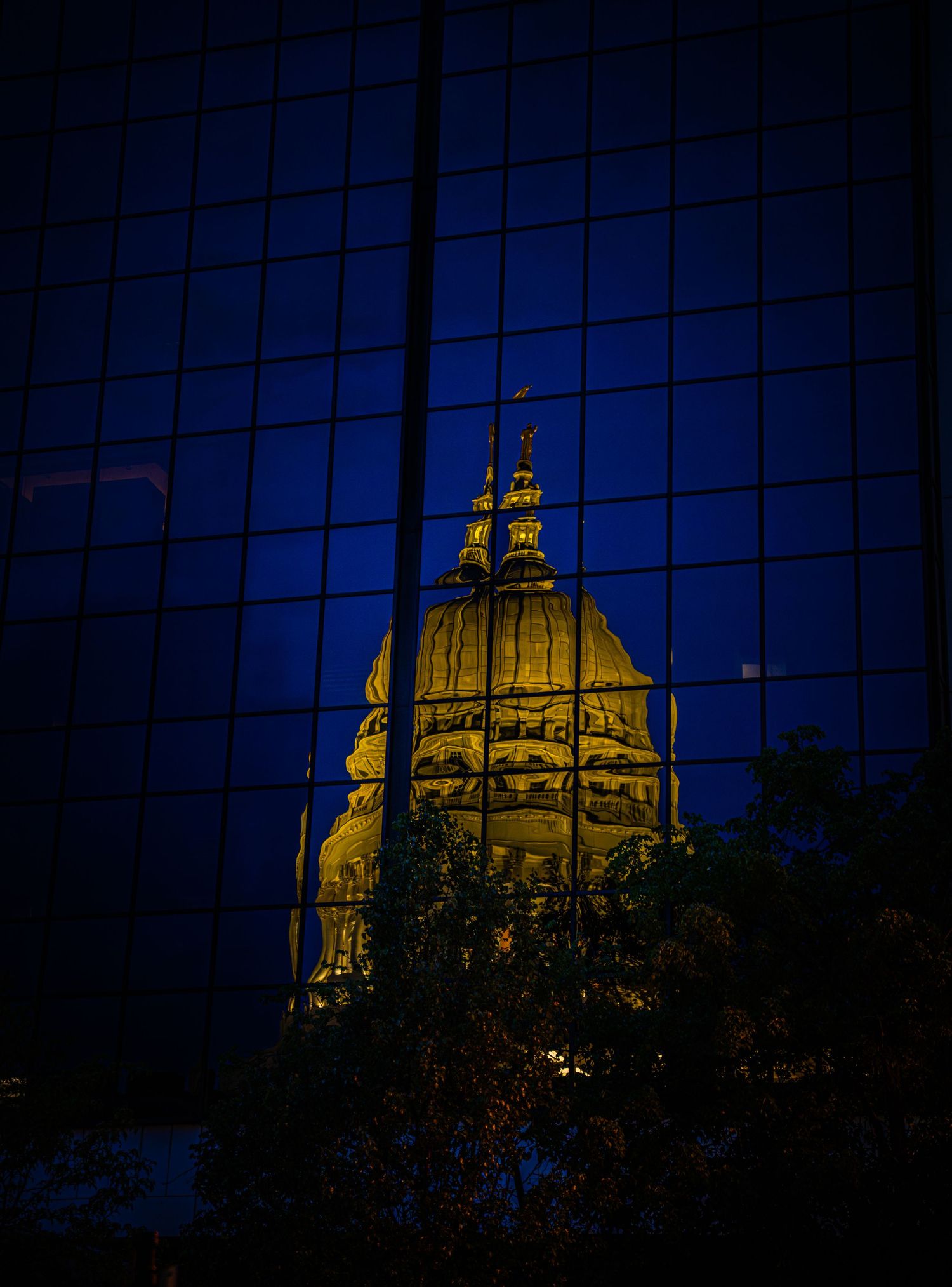 Golden illuminated capitol dome reflected in blue glass office building windows at night with trees below.