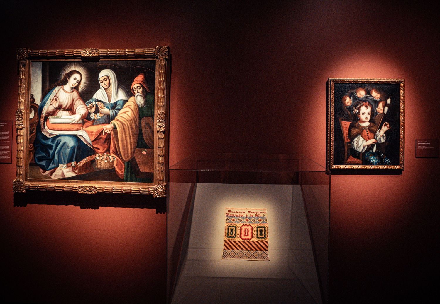 Museum exhibition space showing a series of classical paintings mounted on deep red walls with spotlighting.