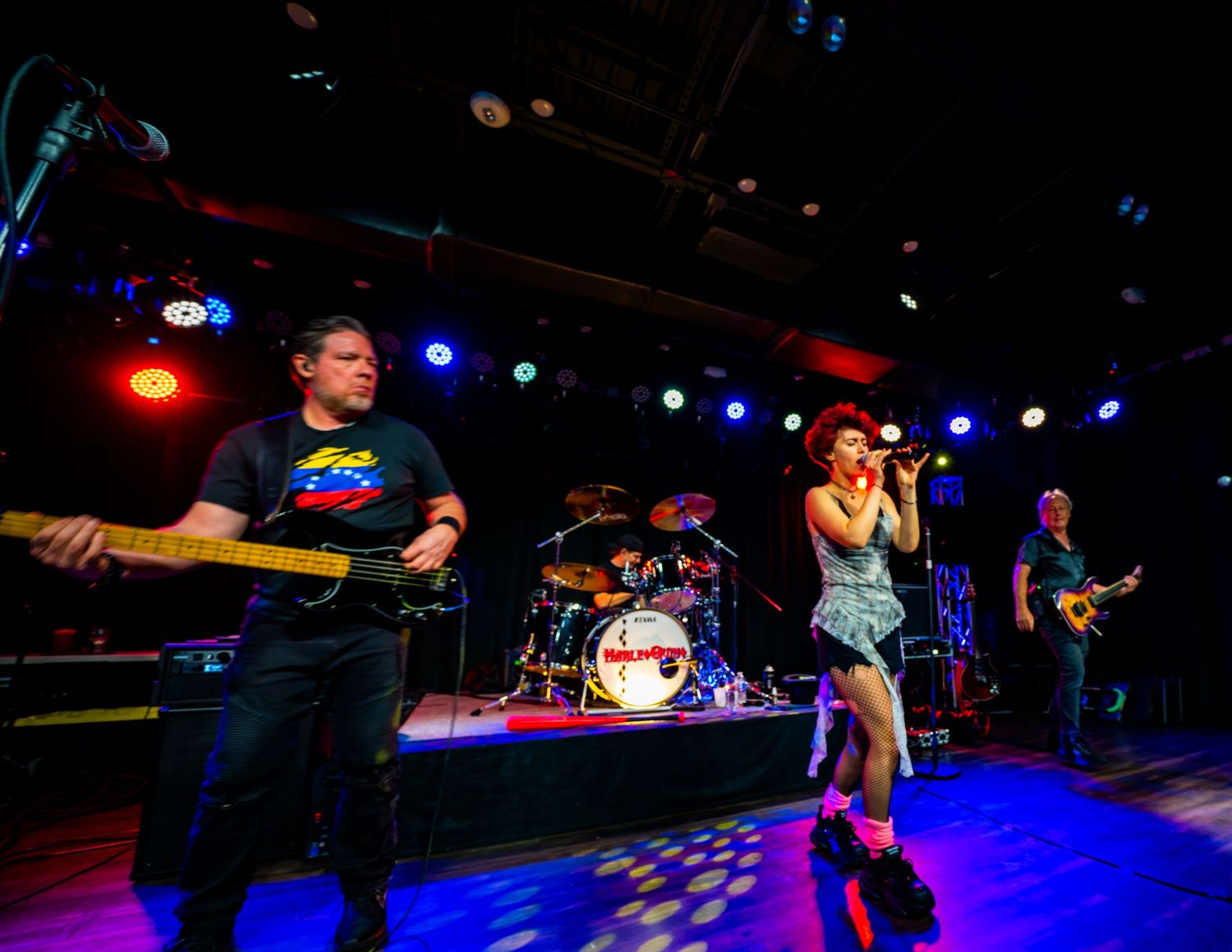 Live rock band performing on stage with colorful lights, guitarist, drummer, female vocalist, and bassist.