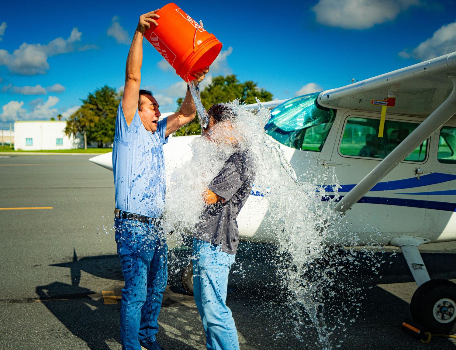 Man dumps orange bucket of ice water on another person near a small plane outdoors, ALS Ice Bucket Challenge.