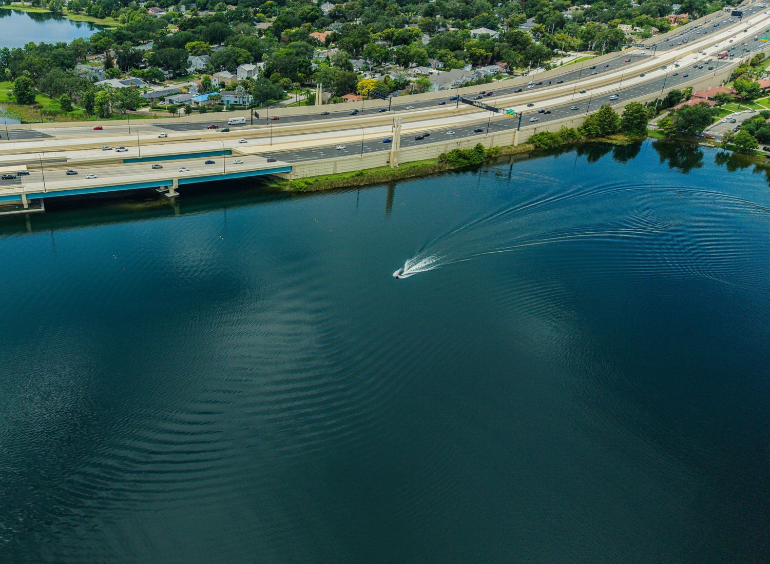 Aerial view of a speedboat leaving rippling wake on deep blue lake near a busy Florida highway bridge.