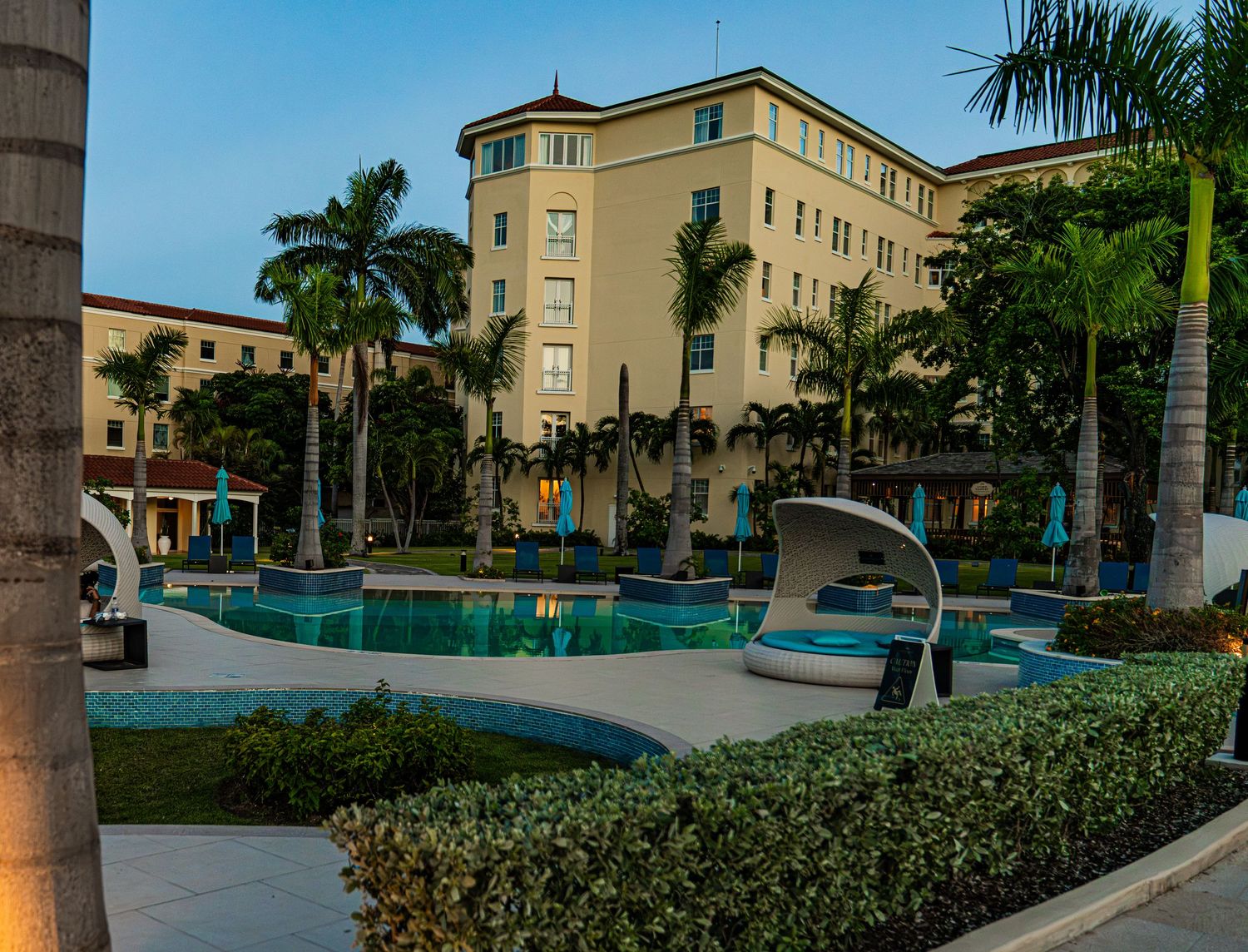 Luxury hotel courtyard with palm trees, blue swimming pool, and elegant lounge chairs at dusk in a tropical setting.
