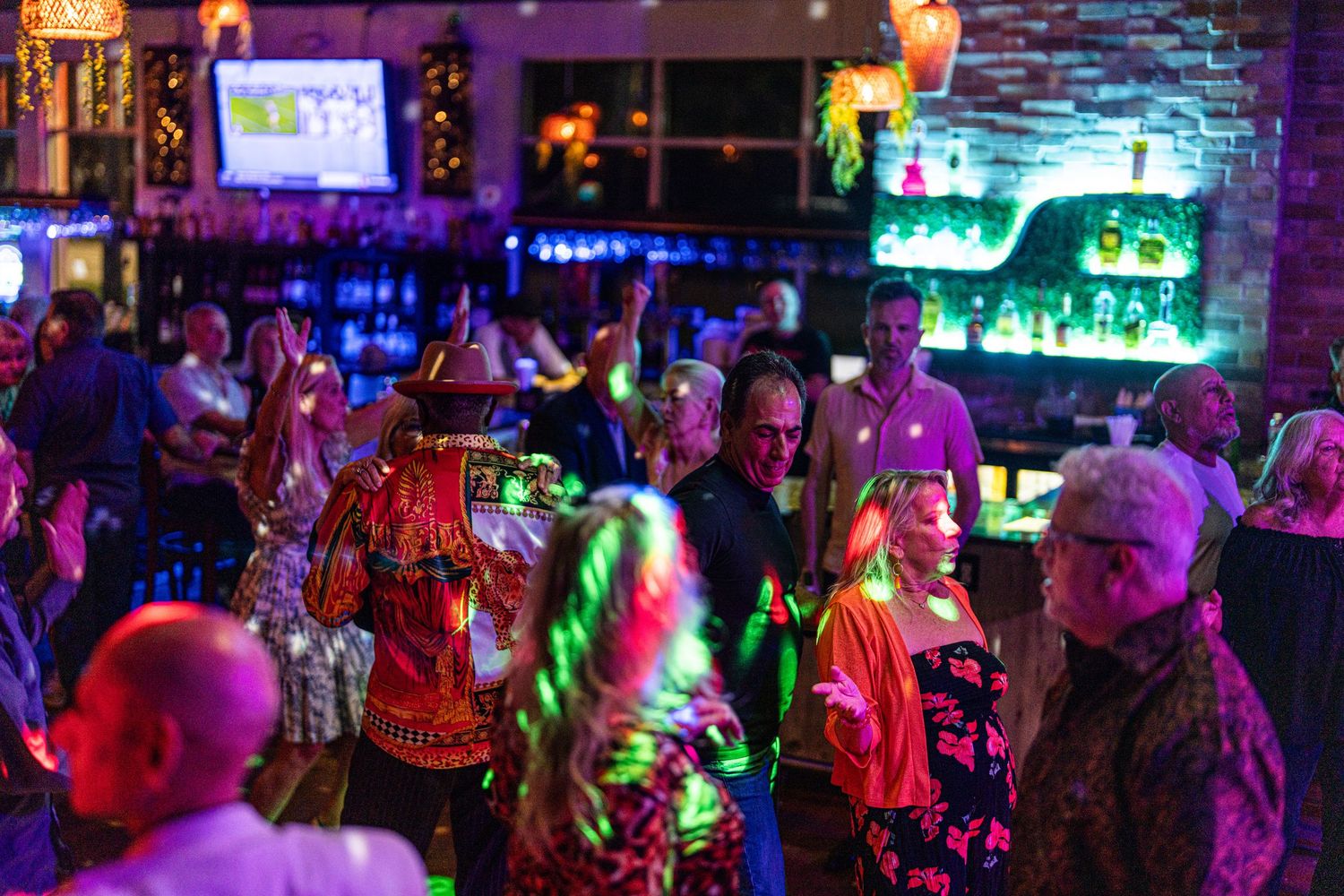 Lively bar scene with colorful lighting, dancing guests, and a vibrant nightlife atmosphere in a crowded venue.