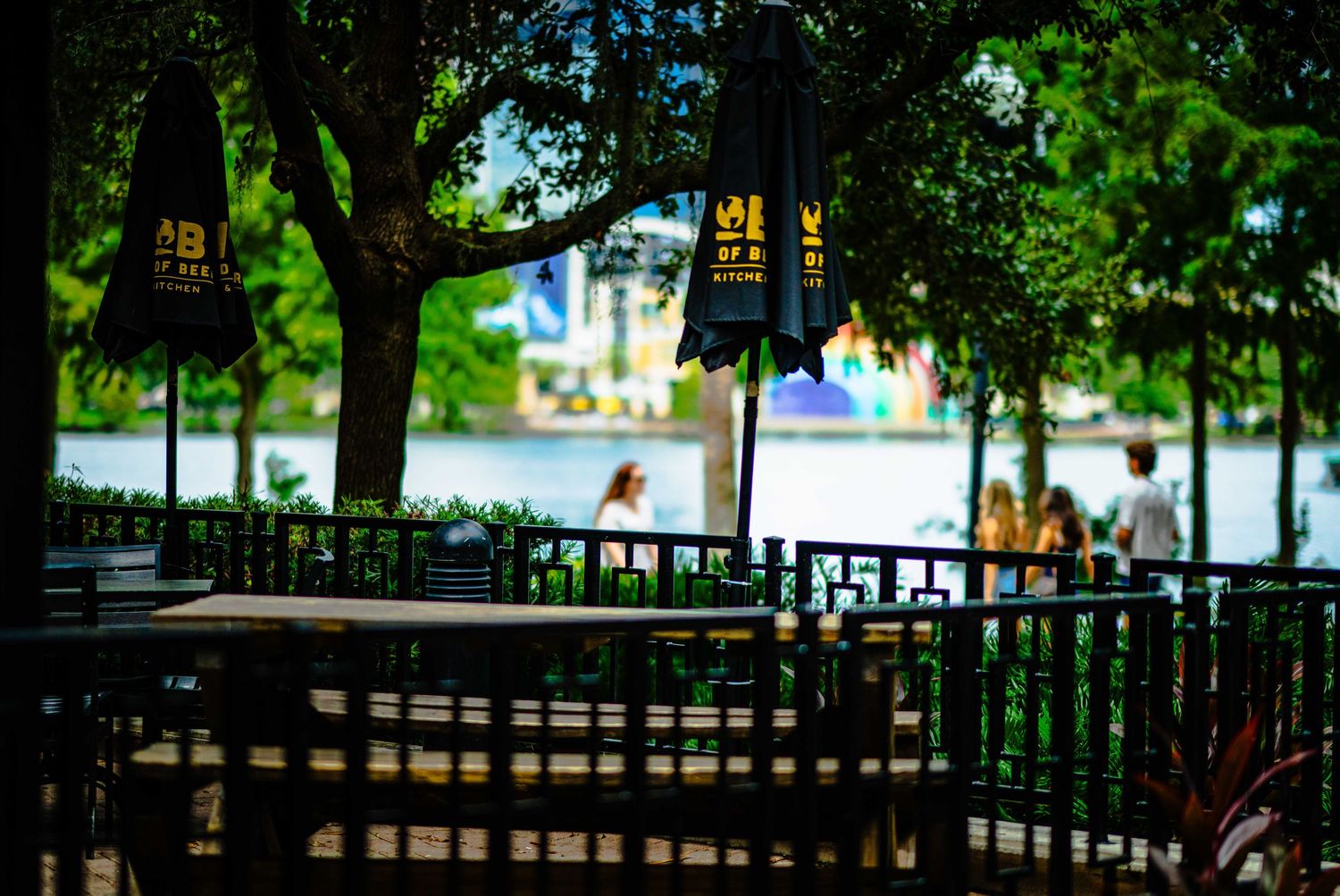 Shaded outdoor patio with wooden tables near a scenic waterfront, surrounded by lush green trees and branded umbrellas.