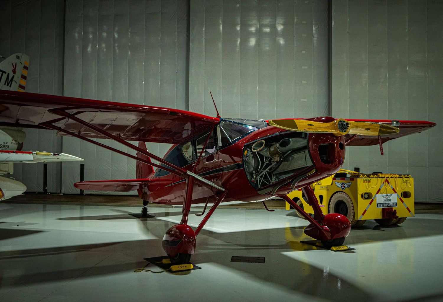 Red vintage small propeller airplane displayed inside a hangar museum with yellow ground equipment nearby.